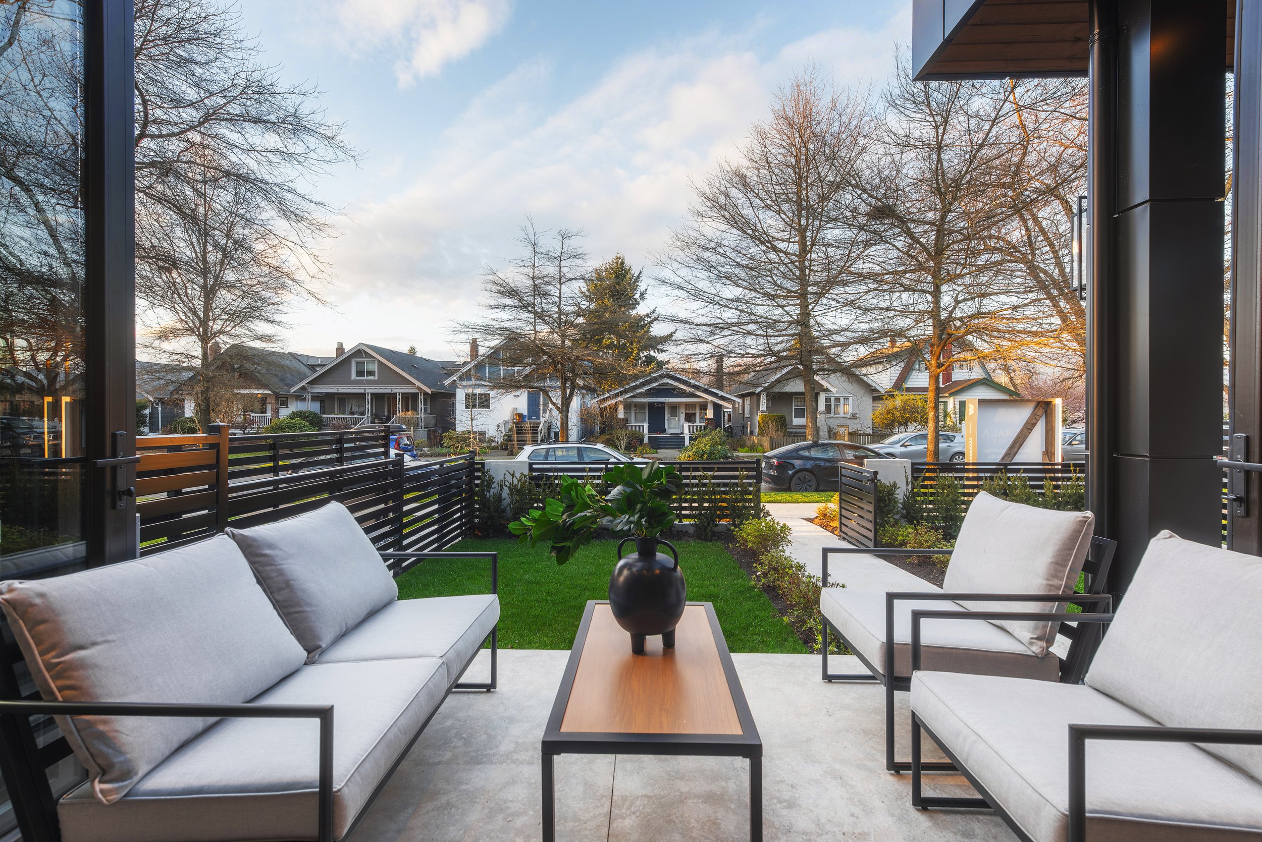 Outdoor patio with modern seating and a small wooden table with a black vase plant, overlooking a green yard and neighboring houses under a cloudy sky.