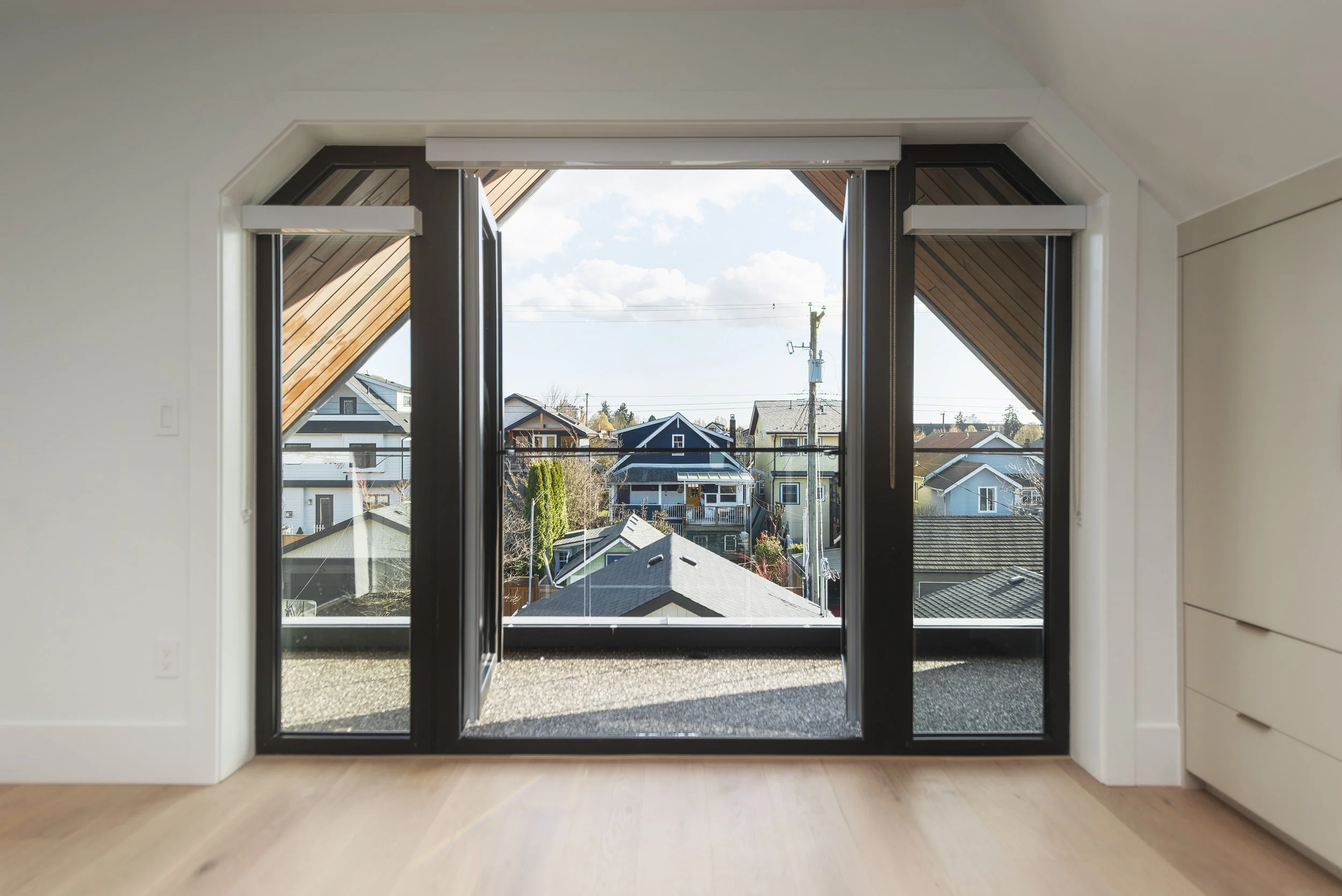 Interior view of a room with large sliding glass doors leading to a small balcony with a view of neighboring houses and power lines under a partly cloudy sky.