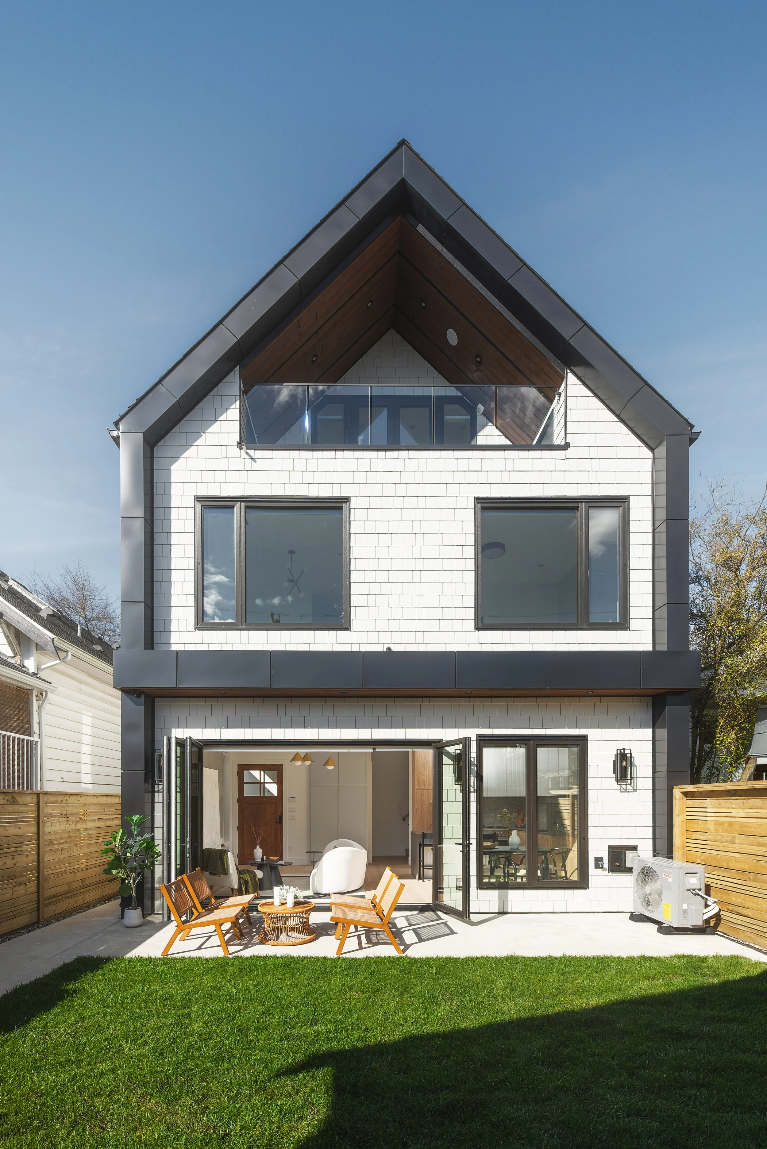 Modern three-story house with white shingles, black trim, large windows, and a front patio with outdoor furniture, surrounded by a green lawn and wooden fencing.