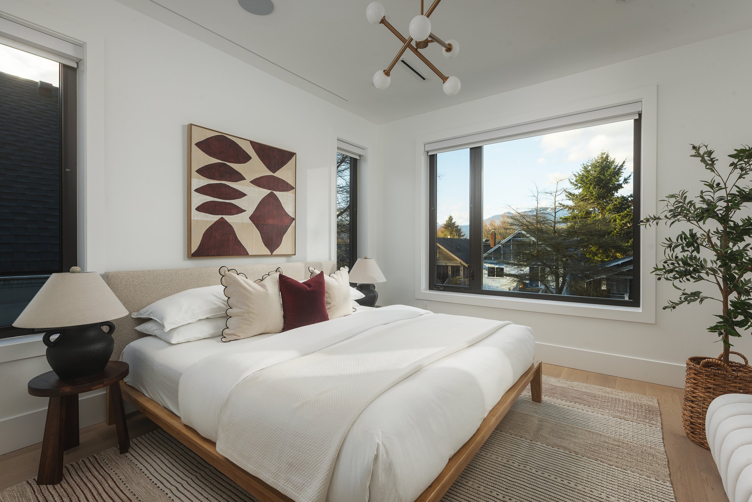 A bedroom with a large bed, two nightstands with lamps, a window showing trees and houses outside, a potted plant in a woven basket, and abstract artwork on the wall.