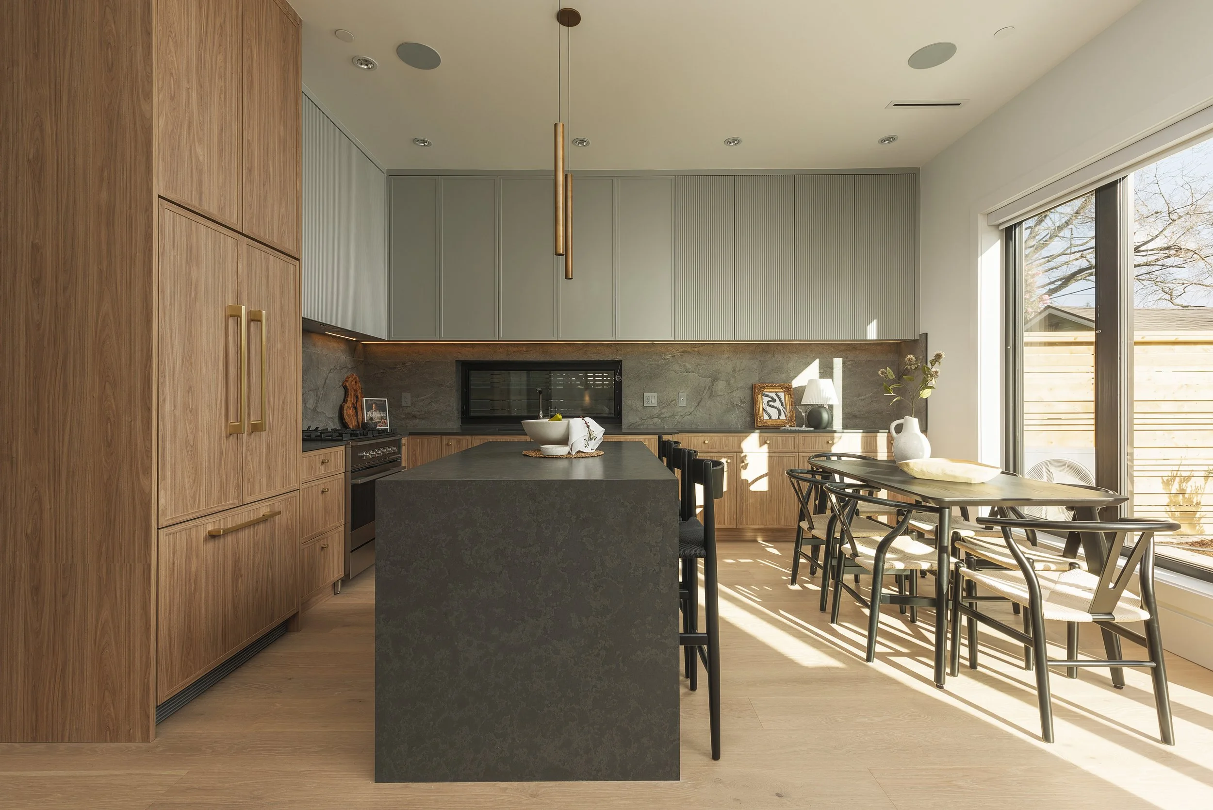 Modern kitchen with wooden cabinets, black island, gray and green upper cabinets, and a dining area by large windows with outdoor view.