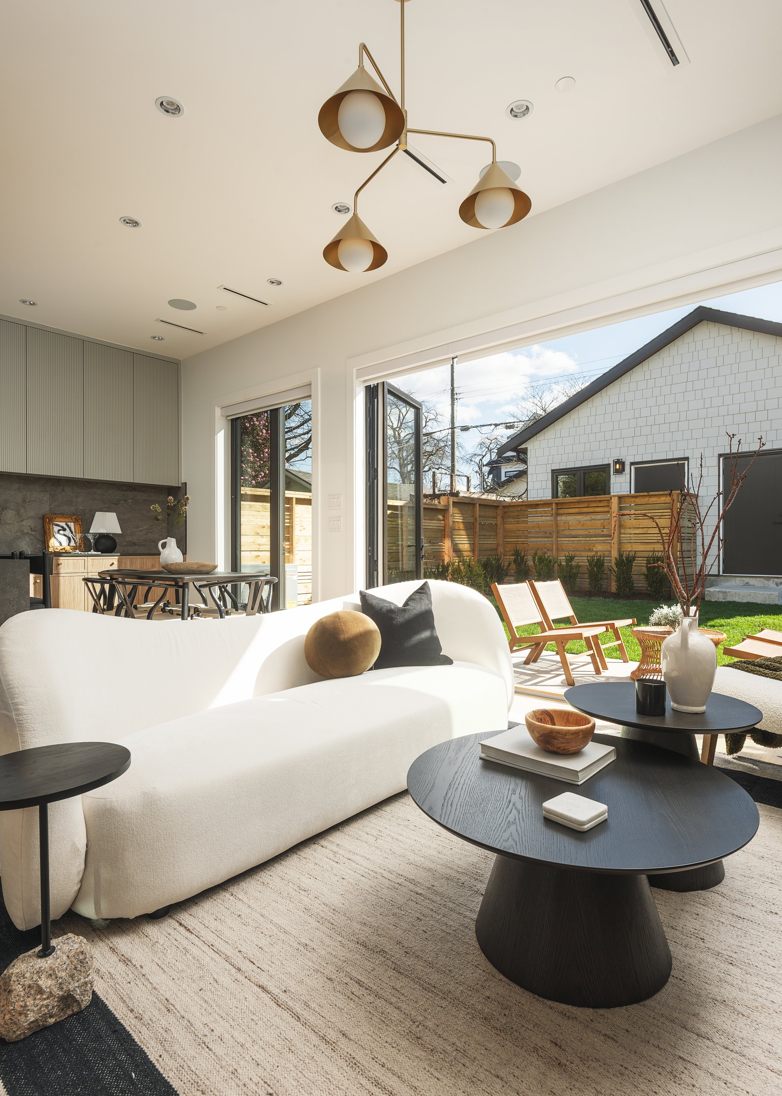 Bright living room with white sofa, black coffee tables, decorative pillows, and large glass sliding doors leading to a backyard with outdoor seating and a fenced yard.