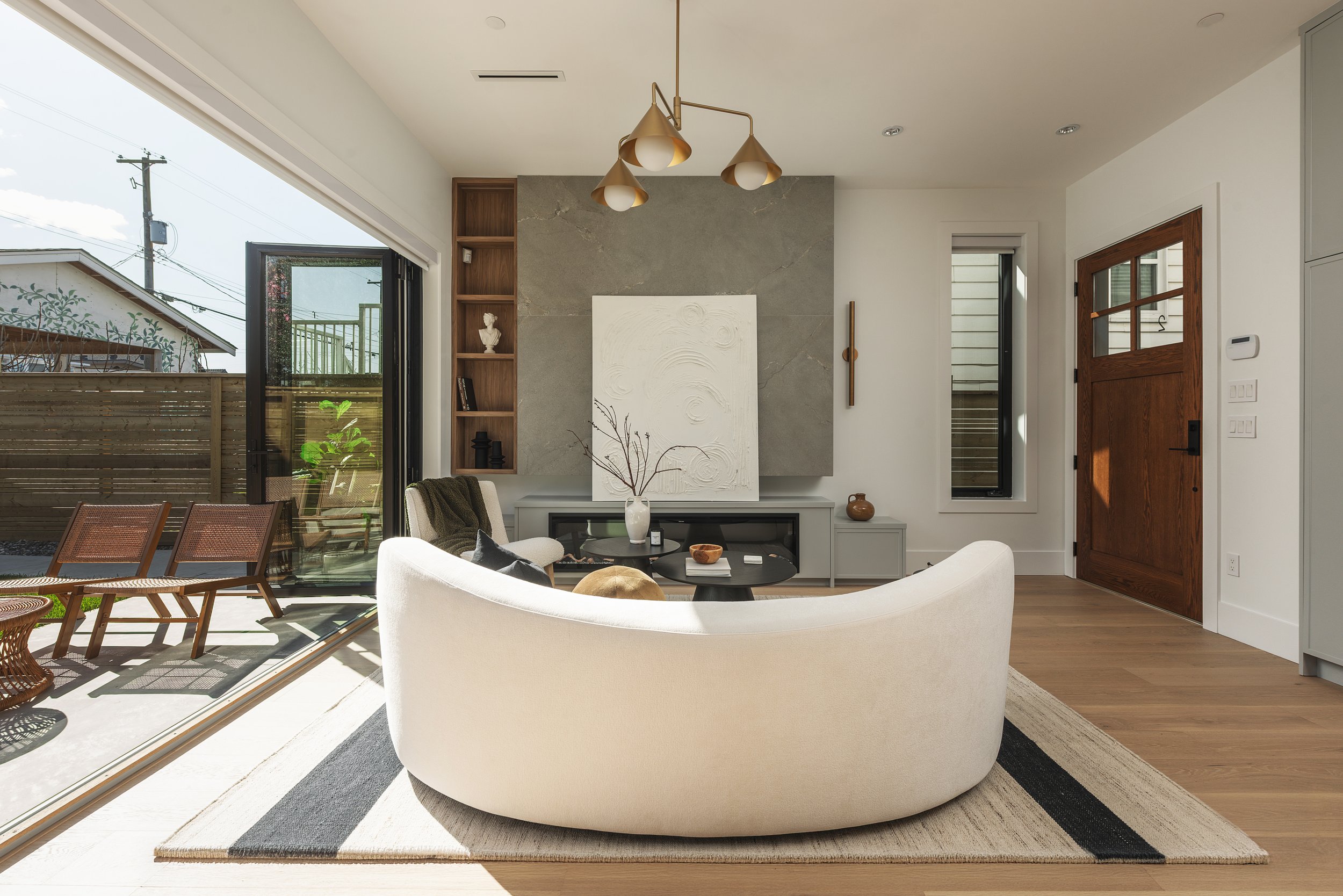 Interior living room with a white curved sofa, black round coffee table with objects, beige rug, fireplace, and artwork on the wall, with large sliding glass door opening to an outdoor patio with chairs and a fence.