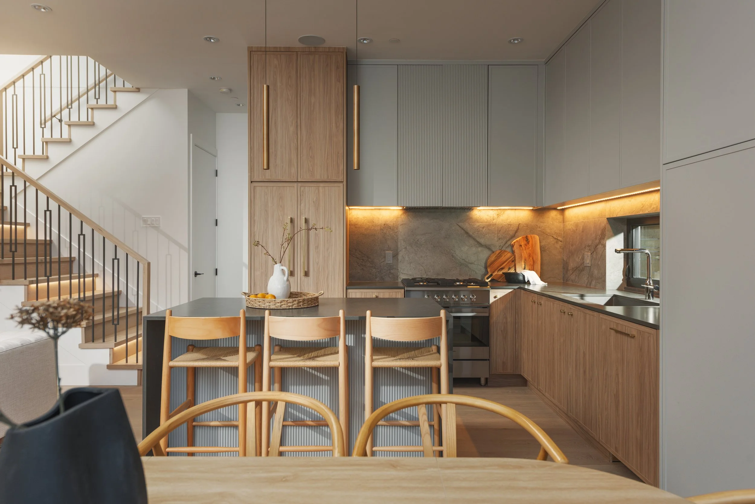 Modern kitchen with wooden cabinets, gray upper cabinets, marble backsplash, black countertop, and a central island with wooden chairs. There is a staircase with wooden steps and black railing on the left.
