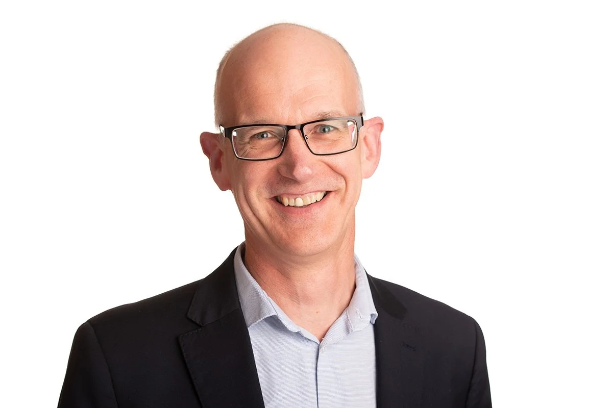 A smiling man with glasses, dressed in a suit, a collared shirt and no tie, standing against a white background.
