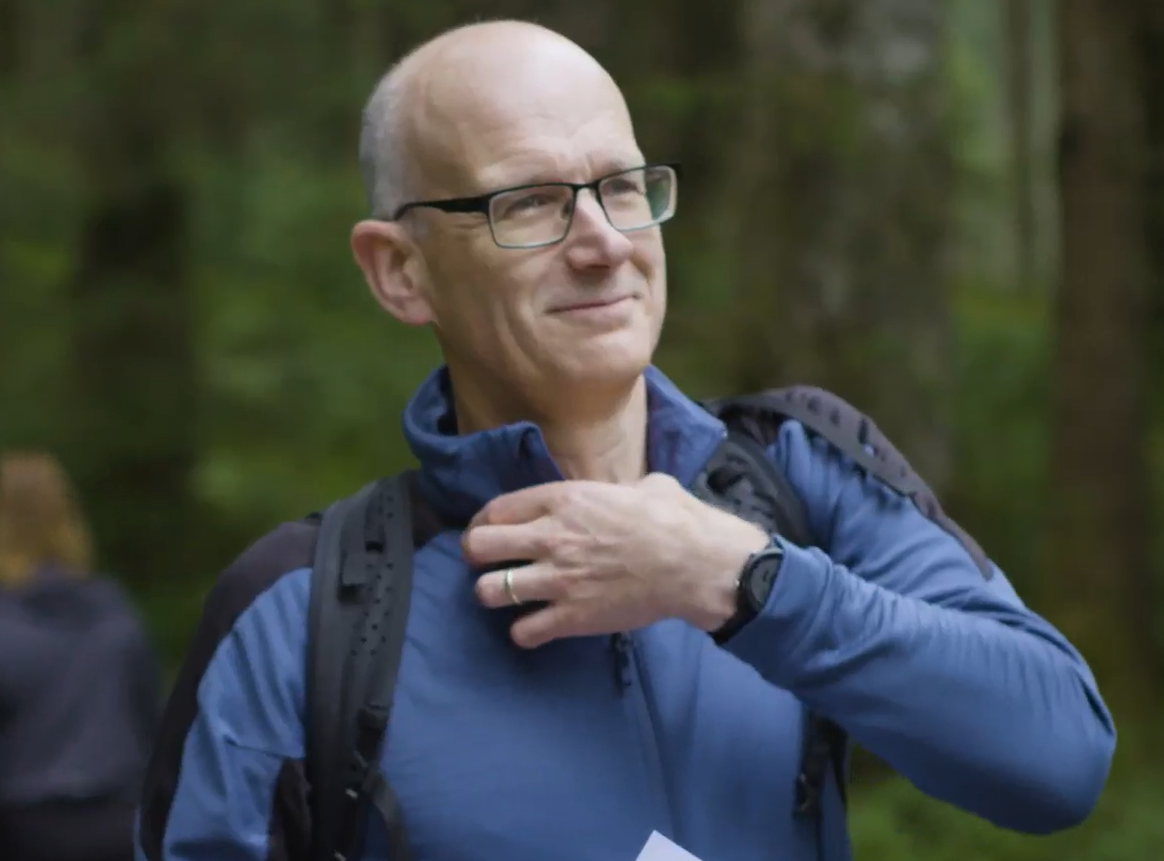 A middle-aged man with glasses and a bald head outdoors in a forest, wearing a blue jacket and a backpack, touching his neck with his leftt hand and smiling.