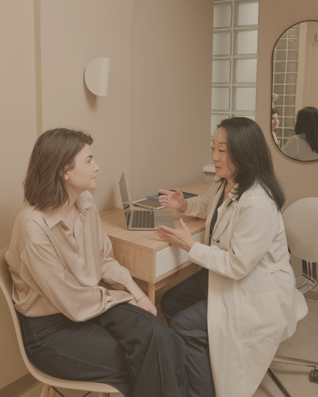 A woman in a beige blouse talks to a doctor in a white coat during a consultation in a medical office.