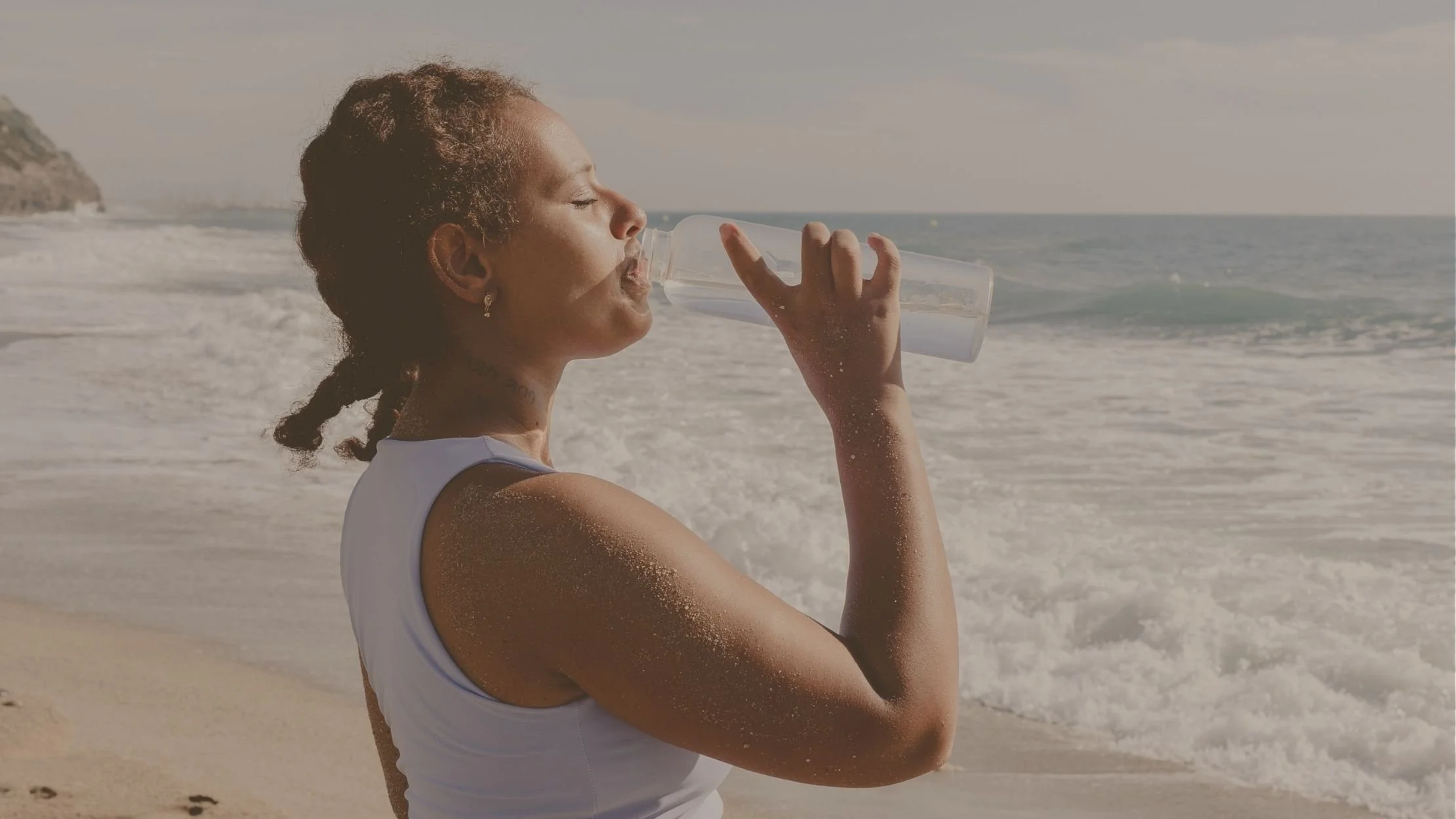 A woman with wet skin and curly hair tied back, wearing a white tank top, drinking water from a clear bottle on a sandy beach with ocean waves and a partly cloudy sky in the background.