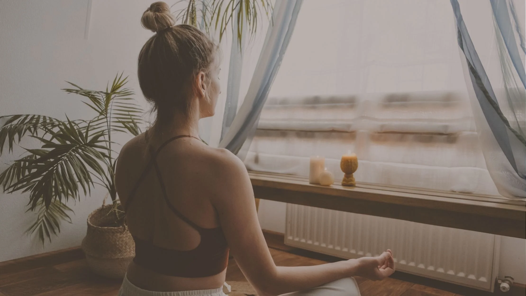 A woman practicing meditation indoors on a wooden floor, sitting cross-legged near a window with sheer curtains, next to a large potted plant, with candles on the windowsill.