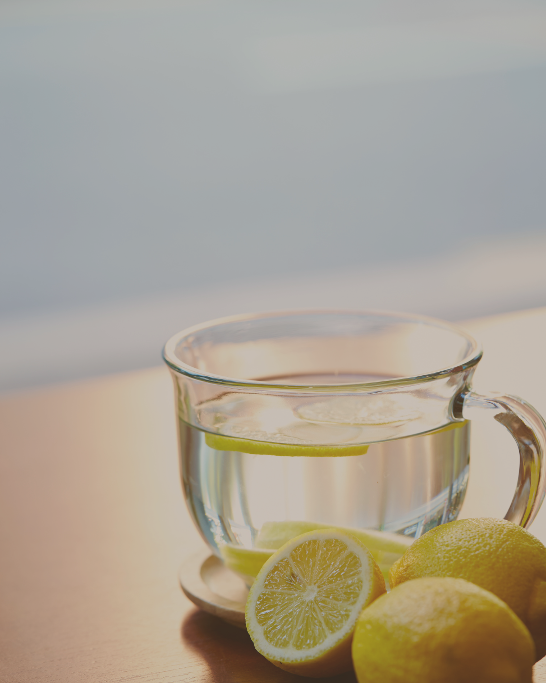 A glass cup of lemon water with lemon slices in and around the glass on a light-colored surface.