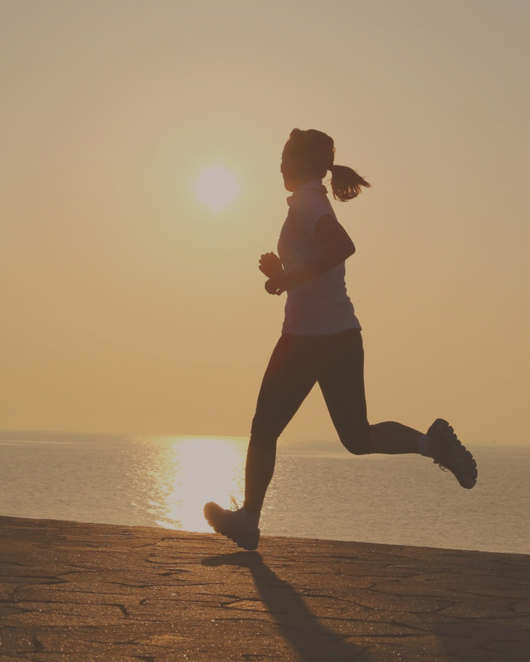 Silhouette of a woman running along a beach during sunset with the sun low in the sky.