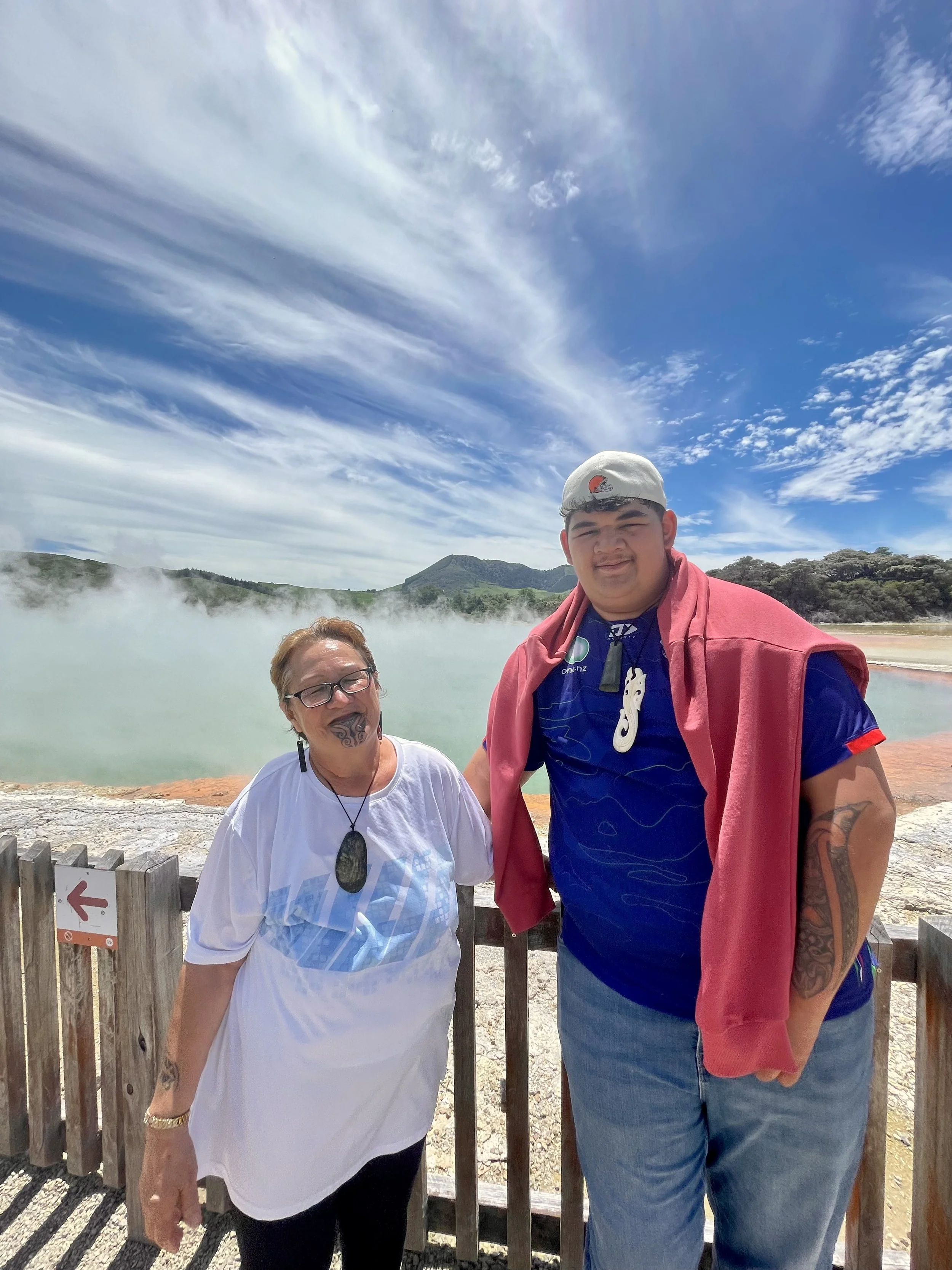 Two people stand in front of a geothermal hot spring with steam rising, blue sky and clouds above, and scenic hills in the background.