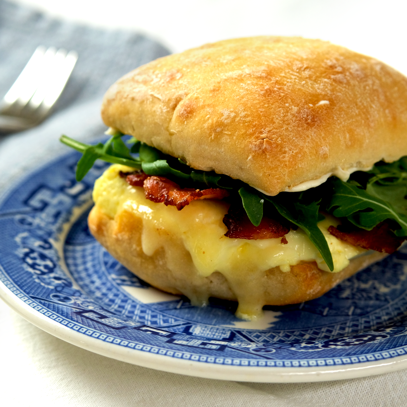 A breakfast sandwich with melted cheese, bacon, scrambled eggs, arugula, on a toasted bun, served on a decorative blue and white plate.
