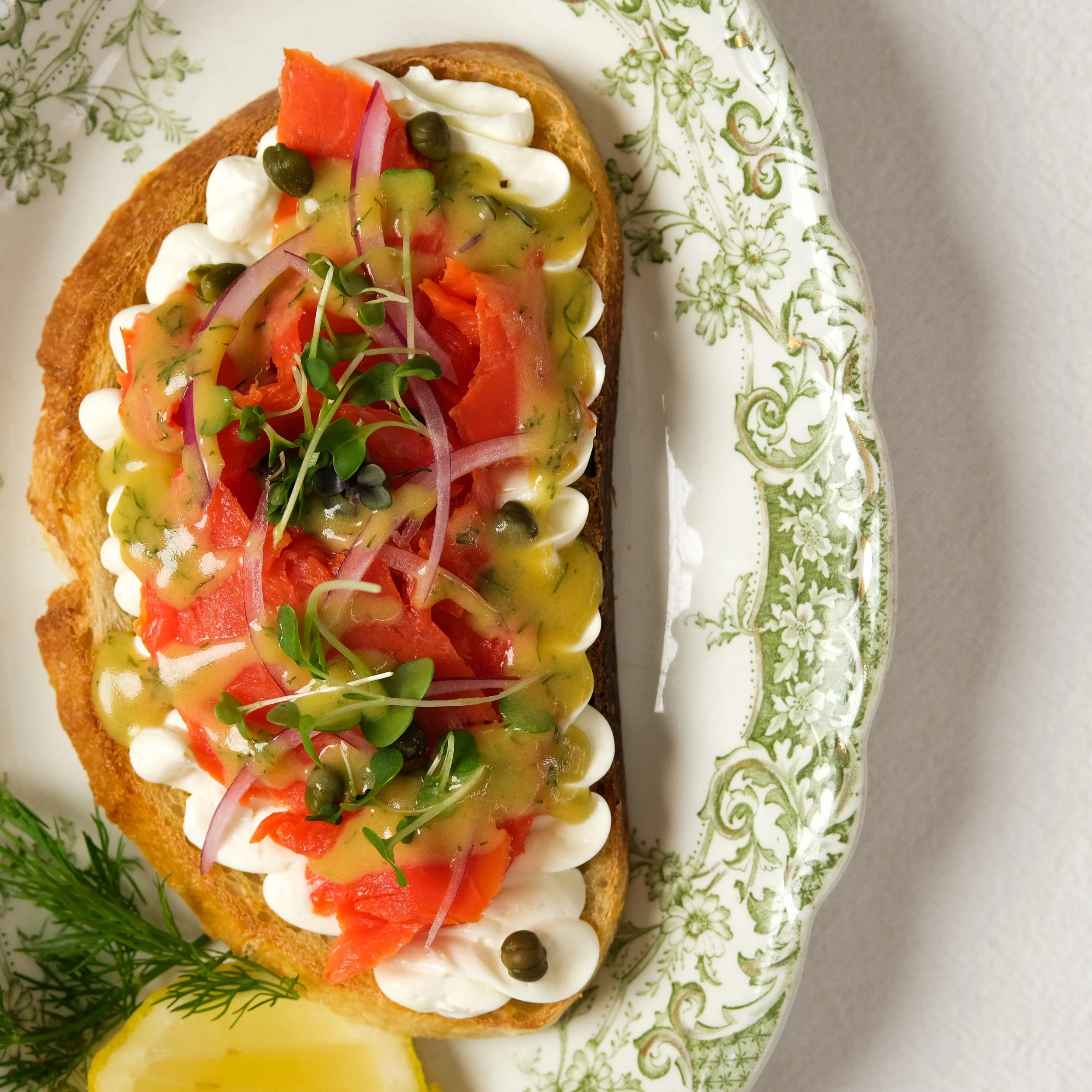 Open-faced sandwich with cream cheese, smoked salmon, red onion, tomato, microgreens, capers, and a drizzle of sauce on a decorative plate with a lemon wedge and garnish.