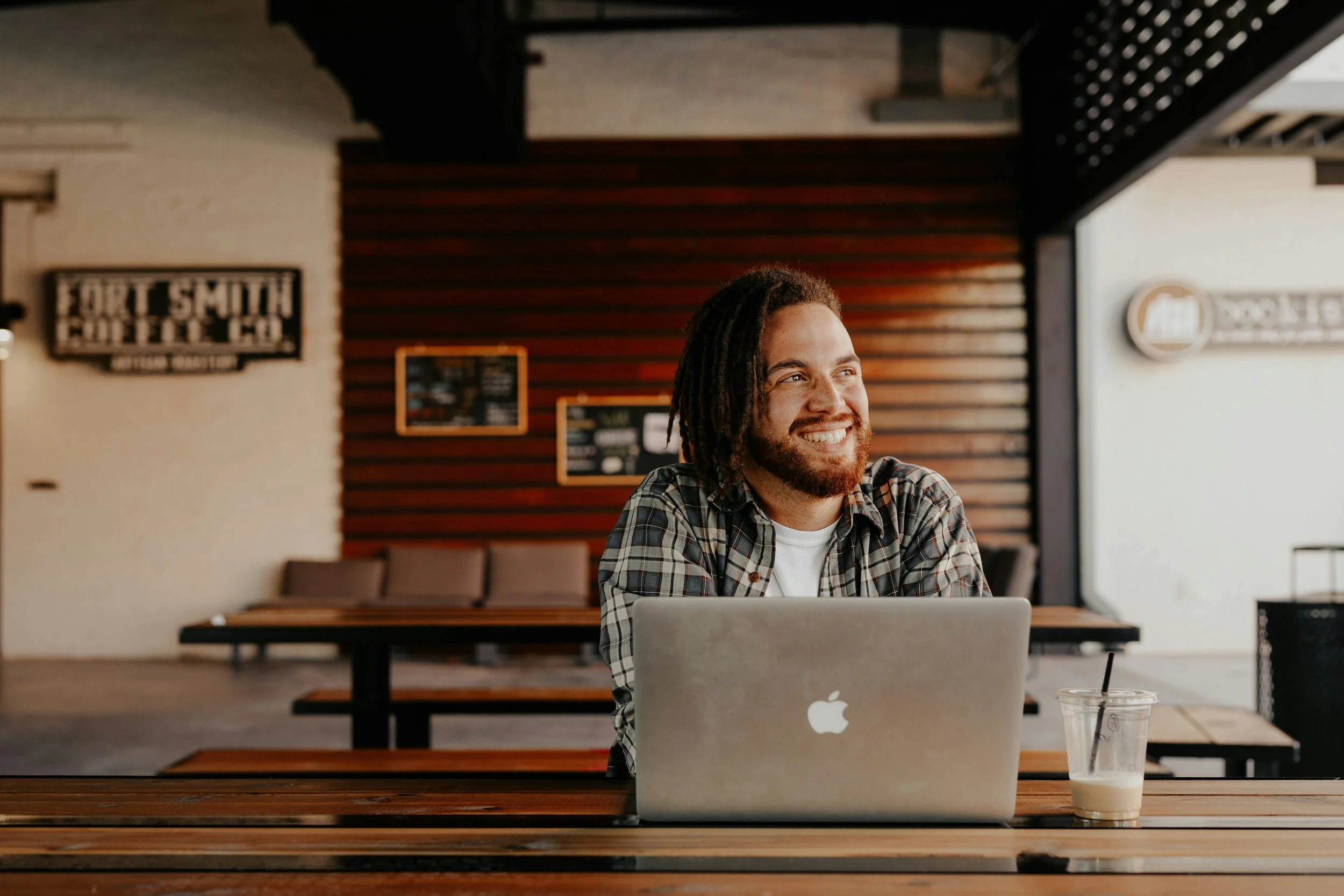 A man with dreadlocks and a beard smiling while sitting at a wooden table with a silver MacBook and iced coffee inside a coffee shop.