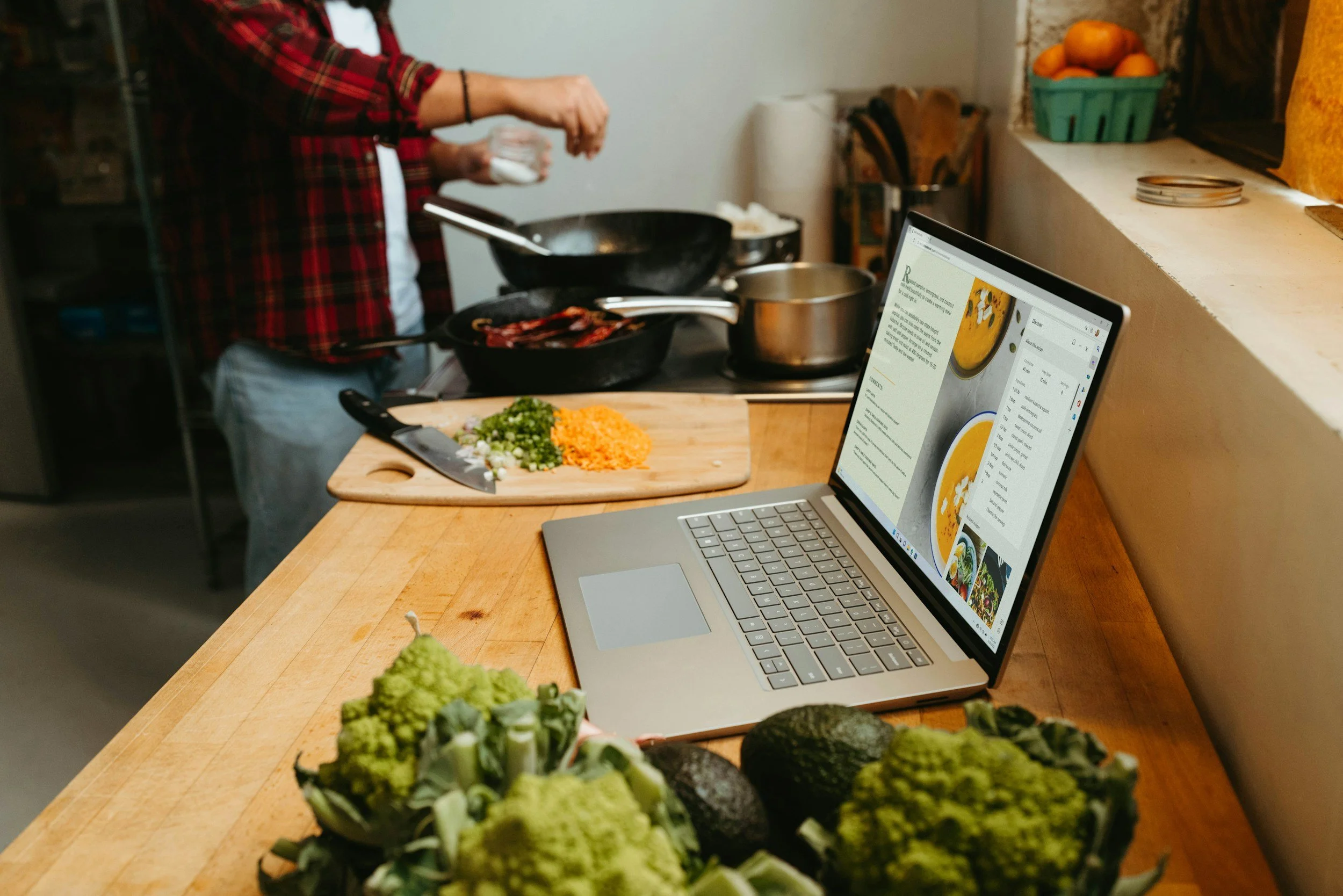 A person cooking in a kitchen with a laptop displaying a recipe. The person is adding salt to a frying pan, with chopped vegetables on a cutting board nearby. There are various pots and kitchen utensils on the counter.