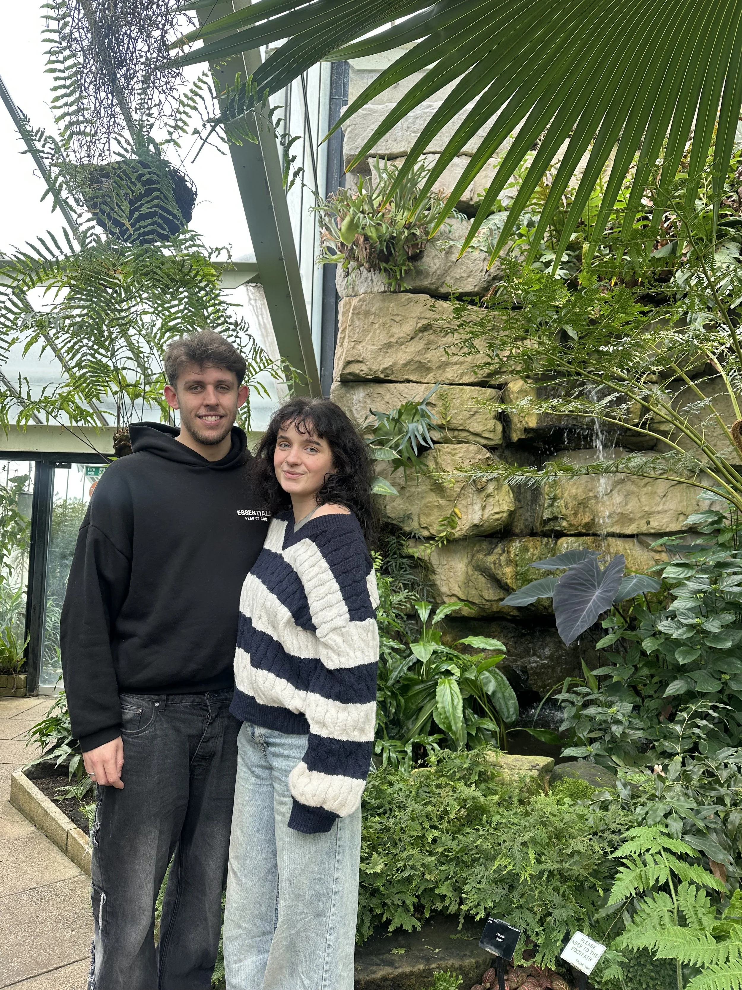 A young man and woman standing close together in a lush indoor greenhouse with a stone waterfall feature, various green plants, and large leaves surrounding them.