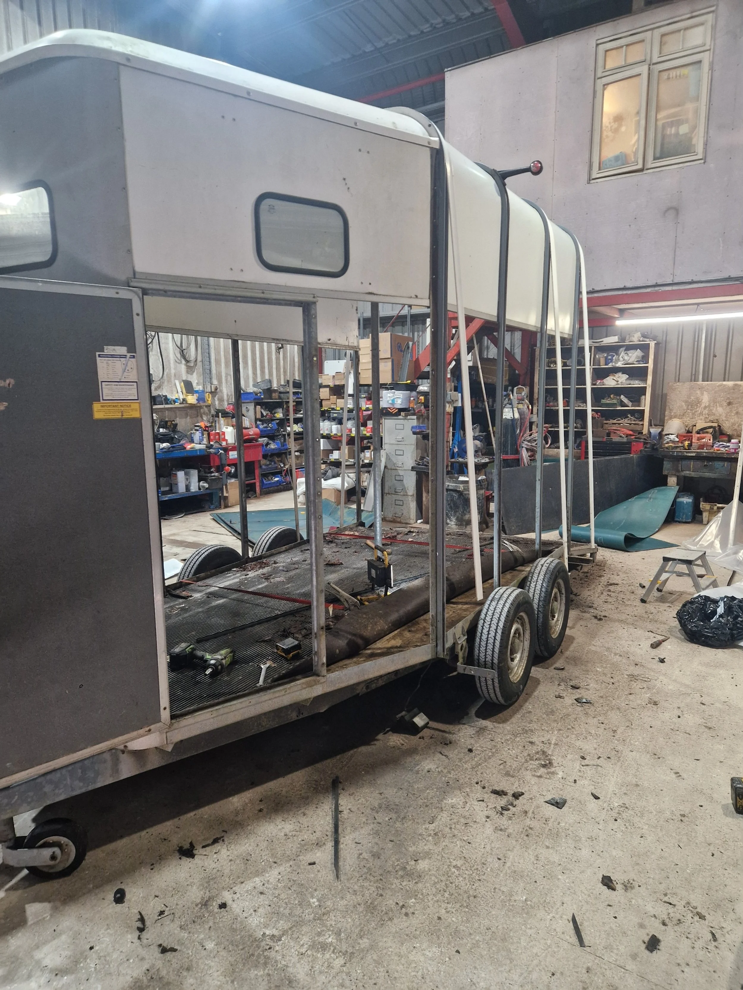A partially assembled utility trailer with a metal frame and wheels, situated in a workshop filled with tools, equipment, and workbenches.