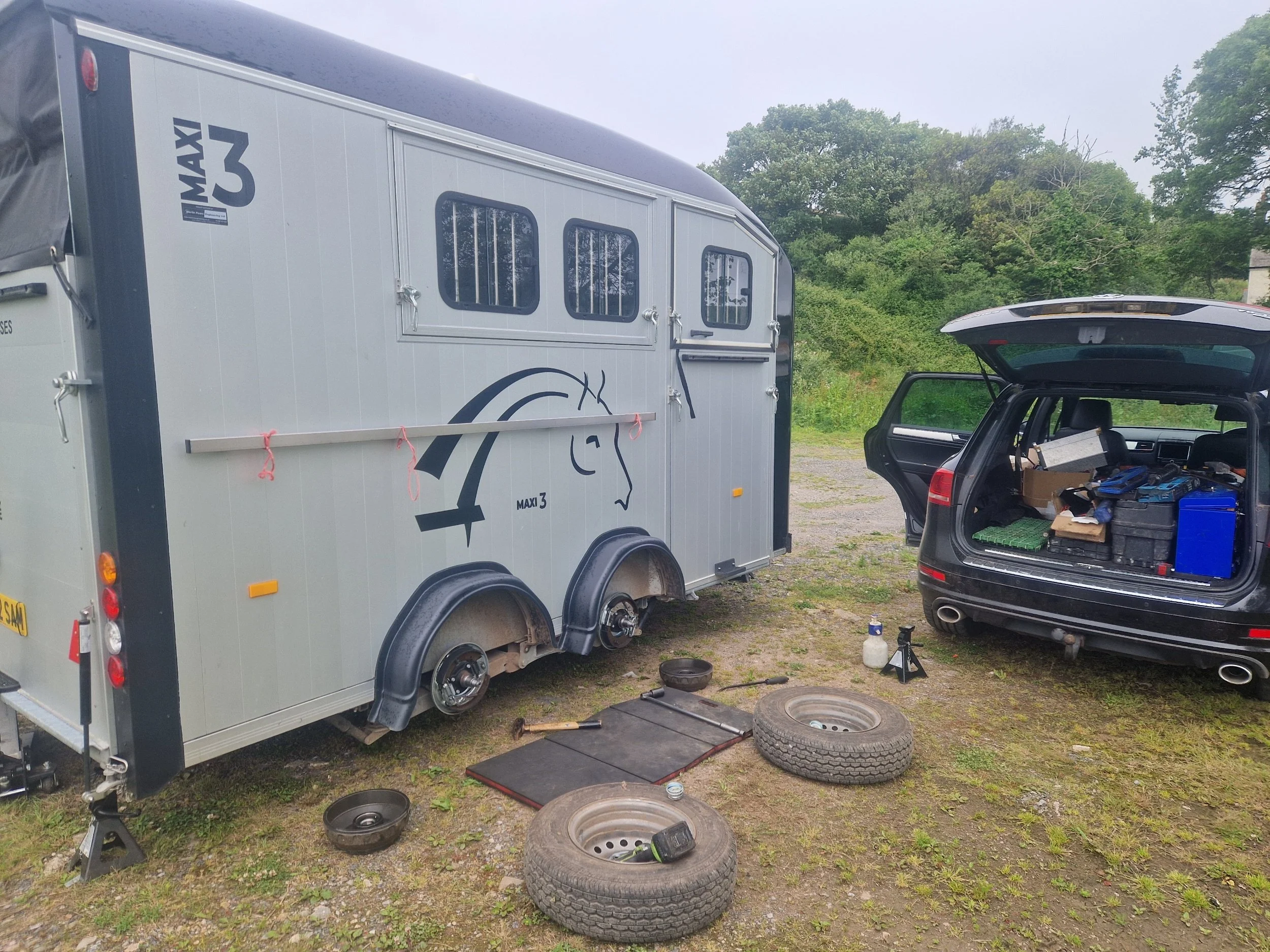 A silver horse trailer labeled 'MAXI 3' parked next to a black SUV with its rear hatch open, showing tools and supplies inside. Two spare tires and various tools are on the ground nearby.