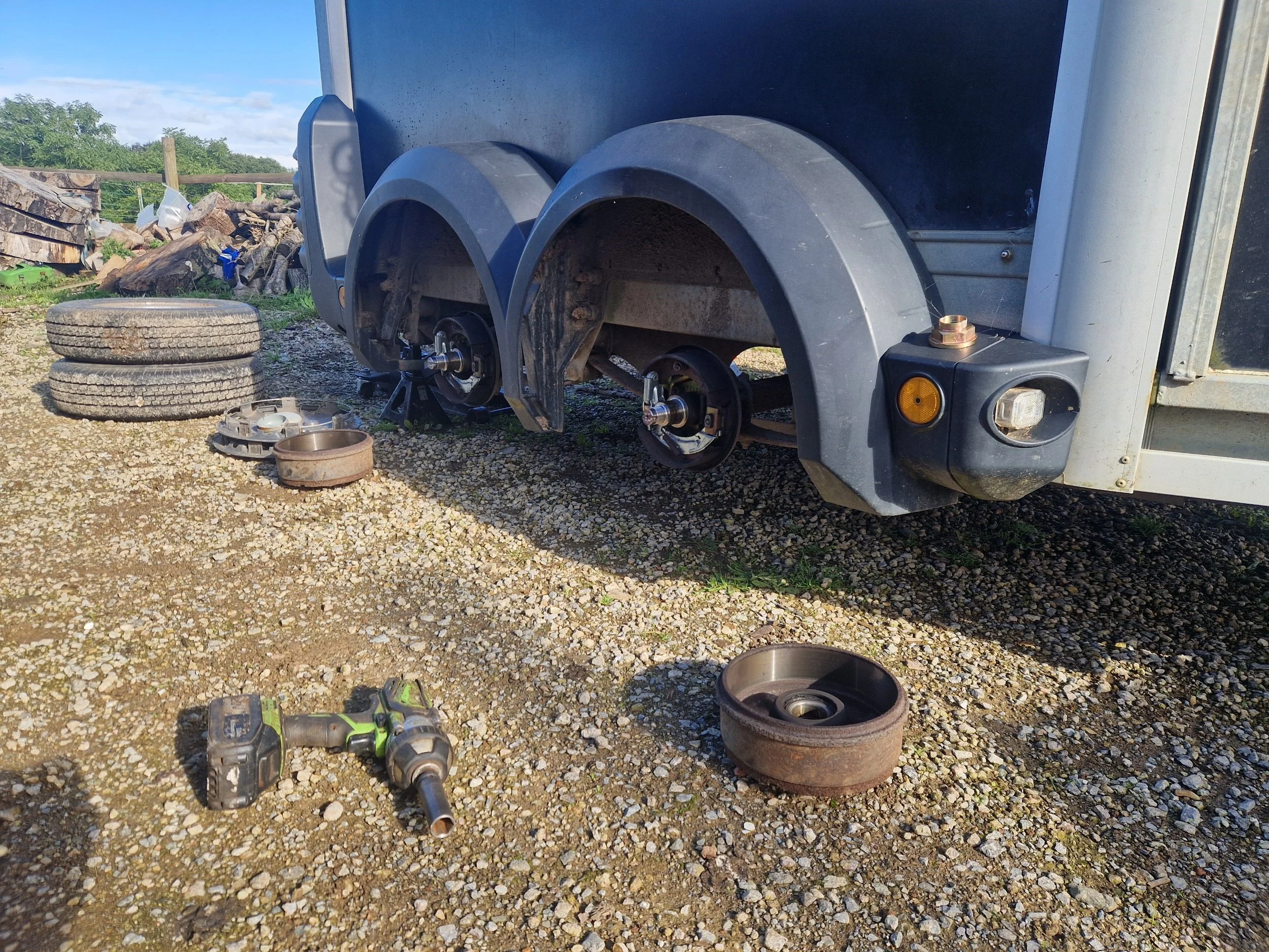 Partially disassembled trailer with removed wheels, truck wheel hubs and tools laid on gravel ground outside.