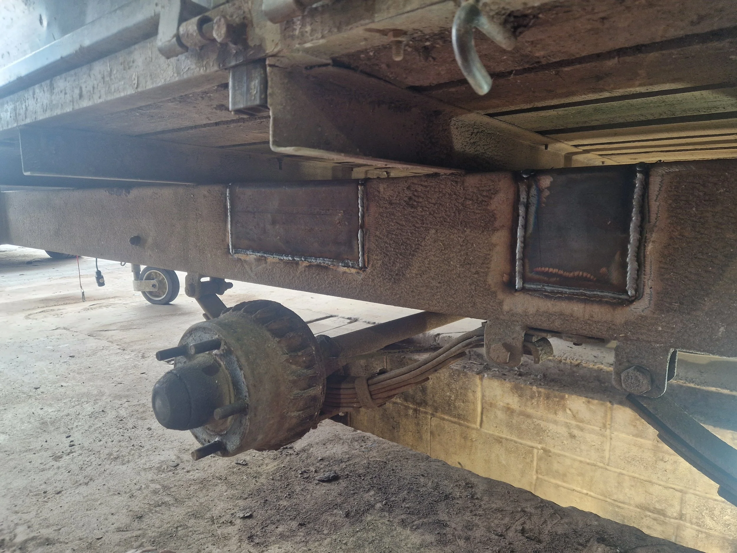 Underneath view of a rusty metal trailer hitch with welded square plates on the side, attached to a weathered trailer frame. The trailer is on a concrete floor with a brick wall in the background.