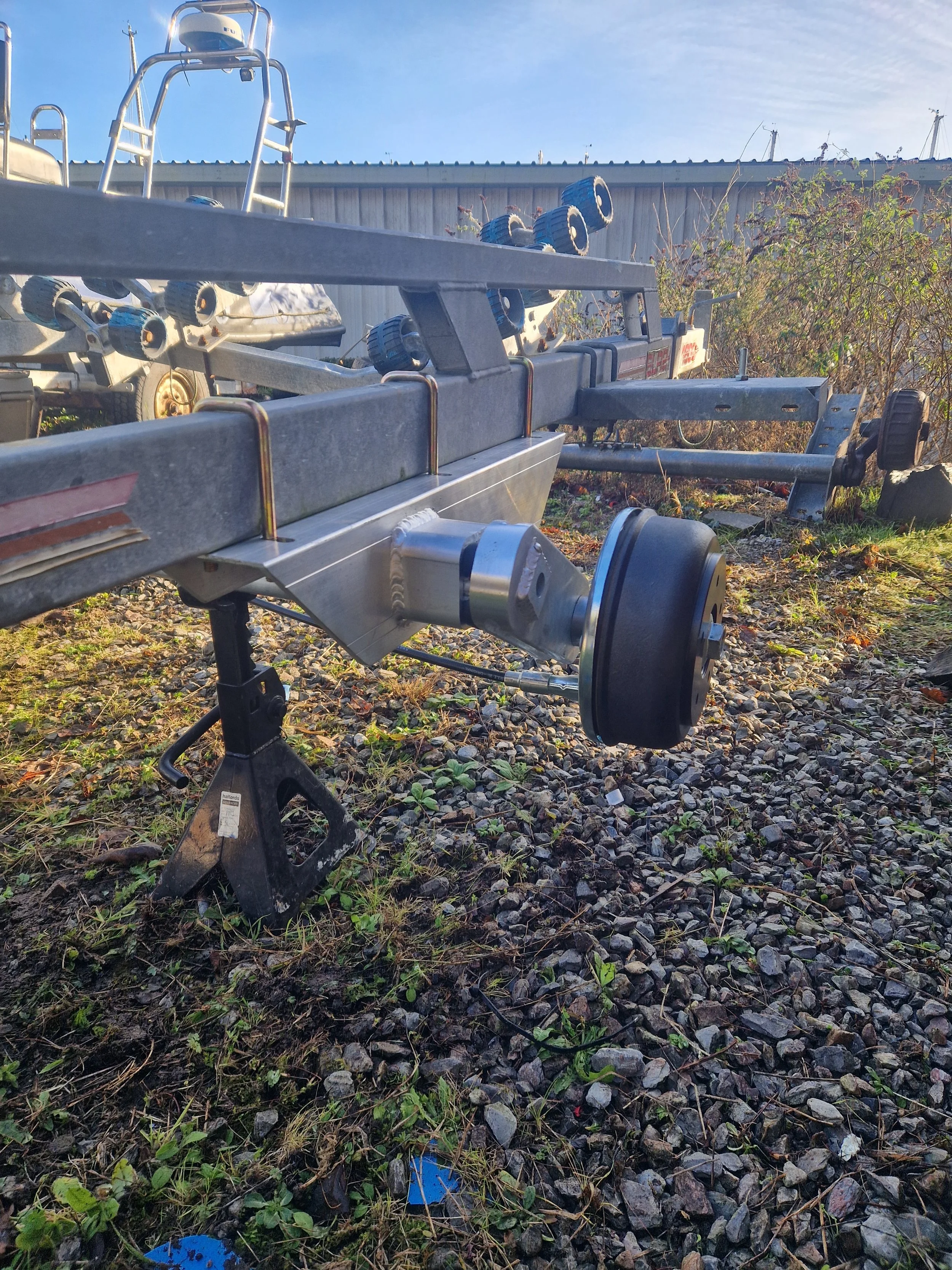 Boat trailer with a black wheel attached, set on uneven gravel ground with some green plants, next to other boat trailers and a metal building, under a clear blue sky.