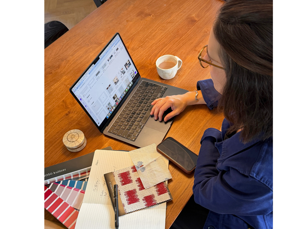 En kvinde med briller sidder ved et træbord, arbejder på en bærbar computer, med en kop te, en smartphone, en farvekortbog, en notesbog med skriver, tekstilprøve og en tekstsamling.»