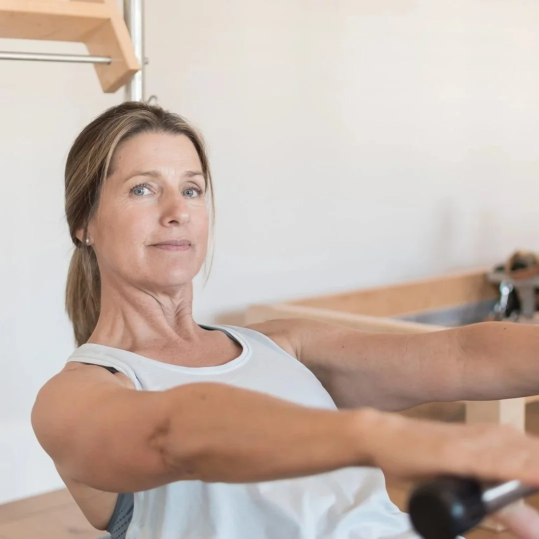 An older woman with brown hair tied back, wearing a white sleeveless athletic top, is exercising with resistance bands in a kitchen or home gym setting. She is pulling the bands forward with her arms extended.