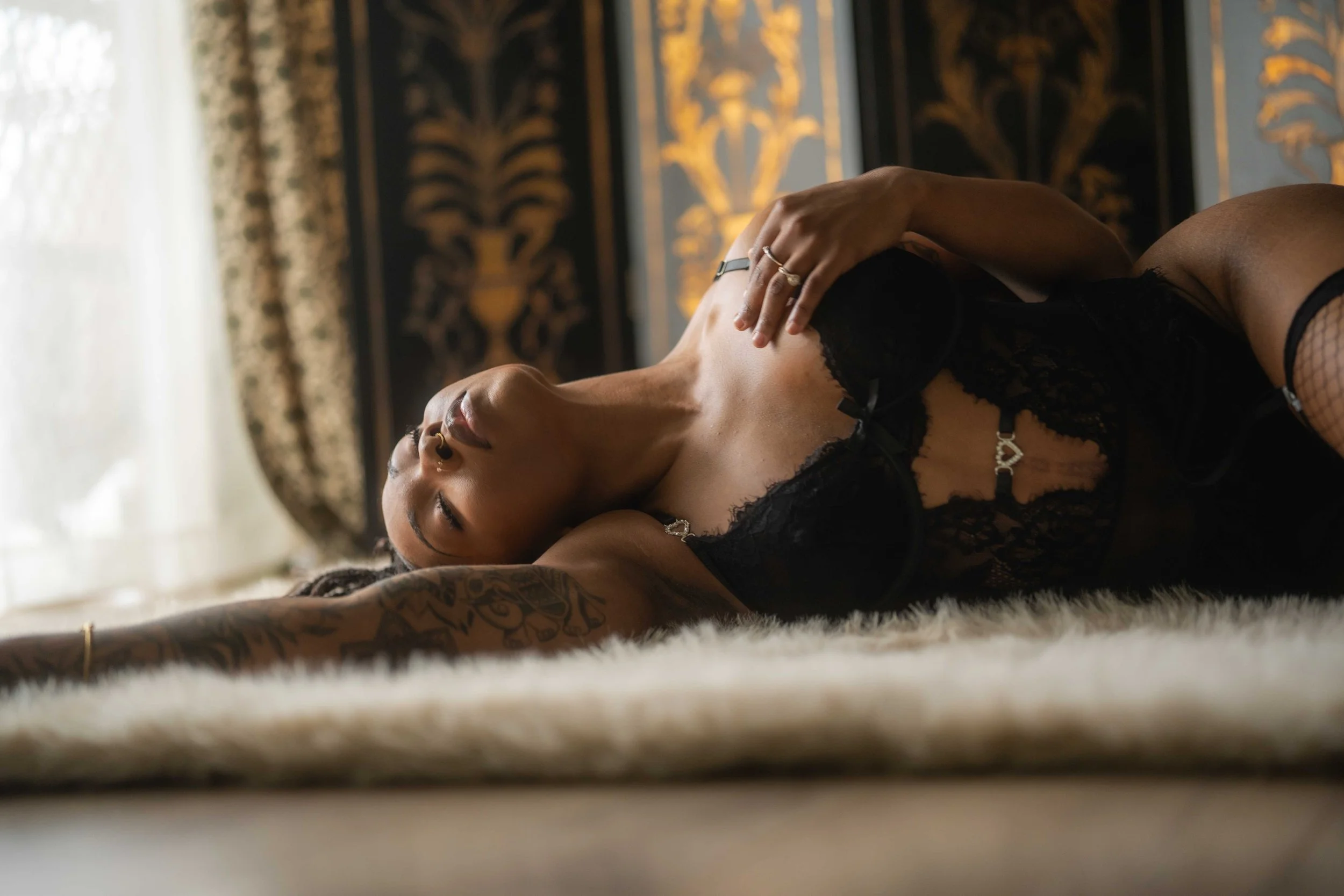 A woman lying on a plush rug on the floor, wearing black lace lingerie, with her eyes closed and one arm extended outward. The background features gold and black ornate wallpaper and a sheer curtain letting in natural light.