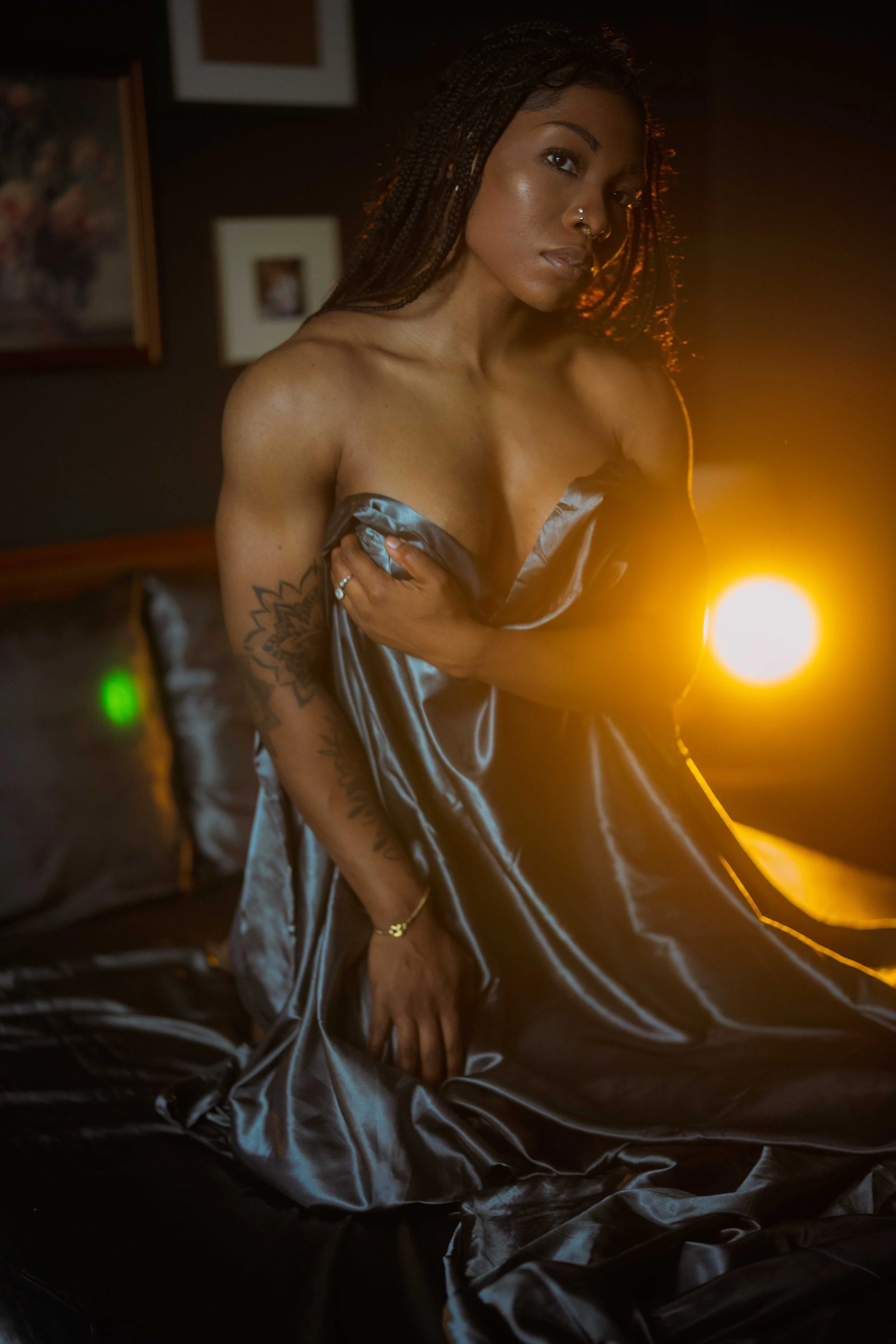 A woman with tattoos on her arm and long braids sits on a bed, covering her chest with a silver satin sheet, illuminated by warm lighting in a dim room with framed pictures on the wall.