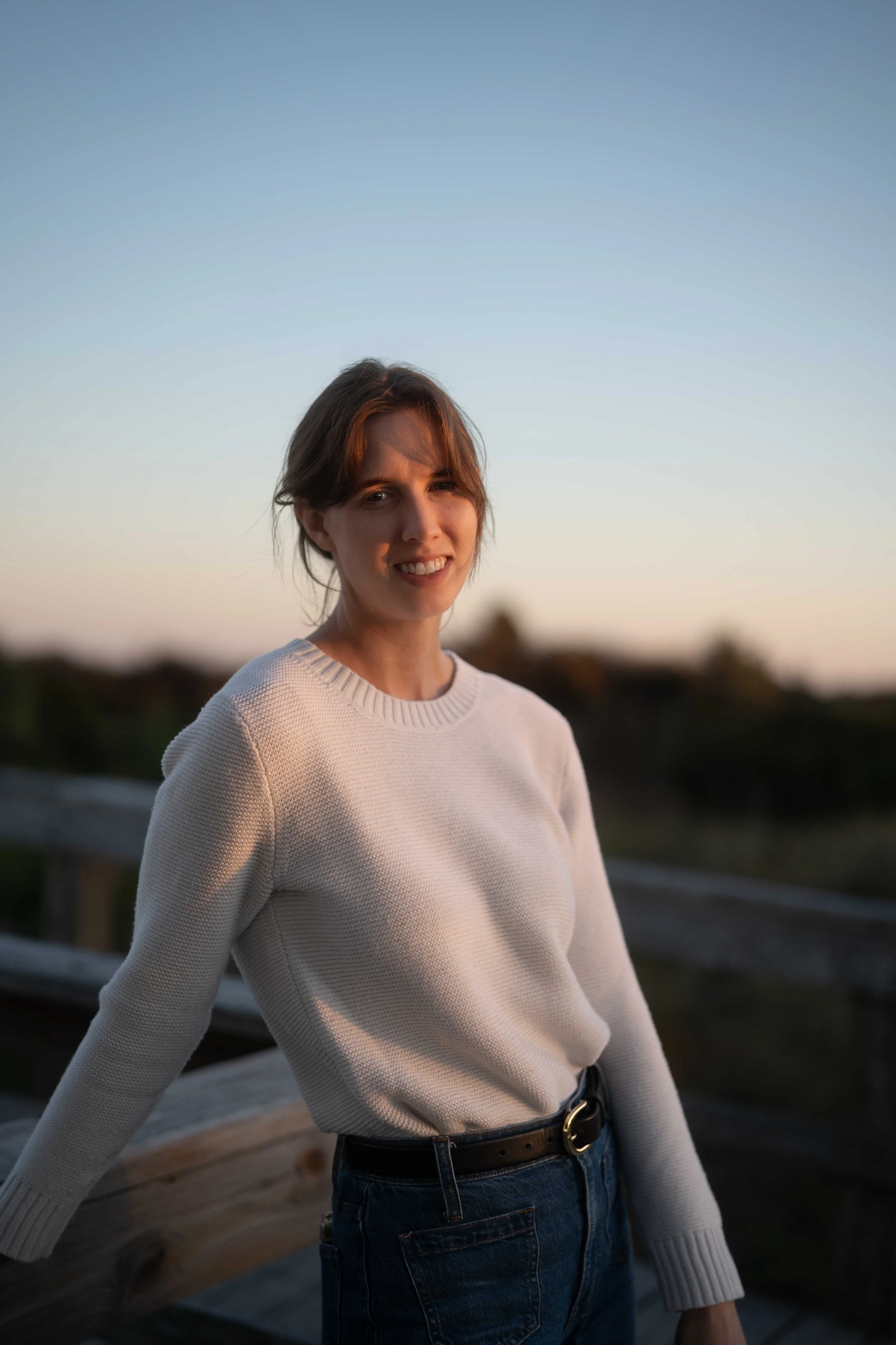 A young woman with shoulder-length brown hair standing outdoors on a wooden bridge at sunset, wearing a white sweater and dark jeans, smiling at the camera.