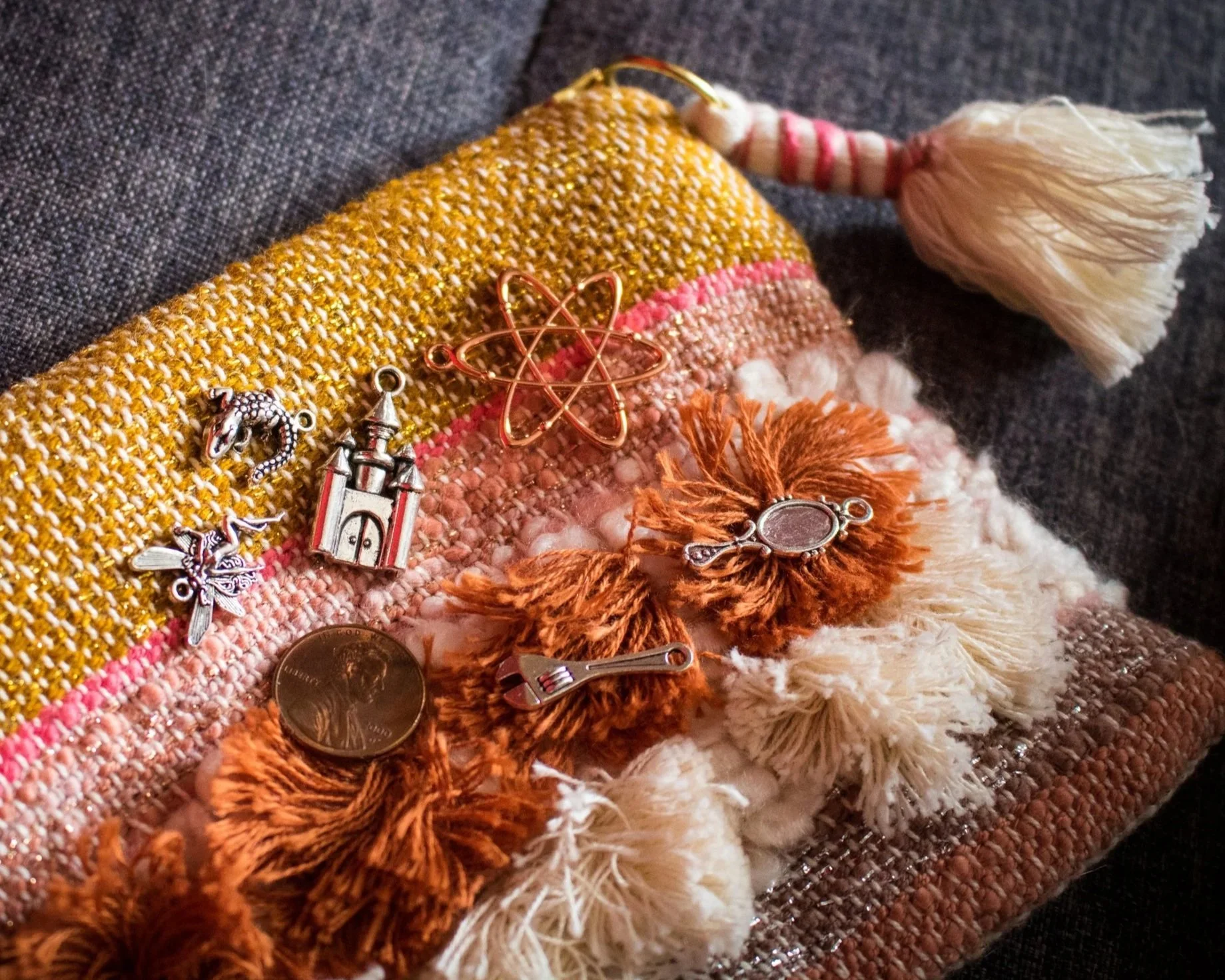 Image of Minerva Siegel's charm casting bag with a few metal charms resting on it. The bag is made of natural fibers and woven with a tassel, and the charms include a small castle and atomic symbol.