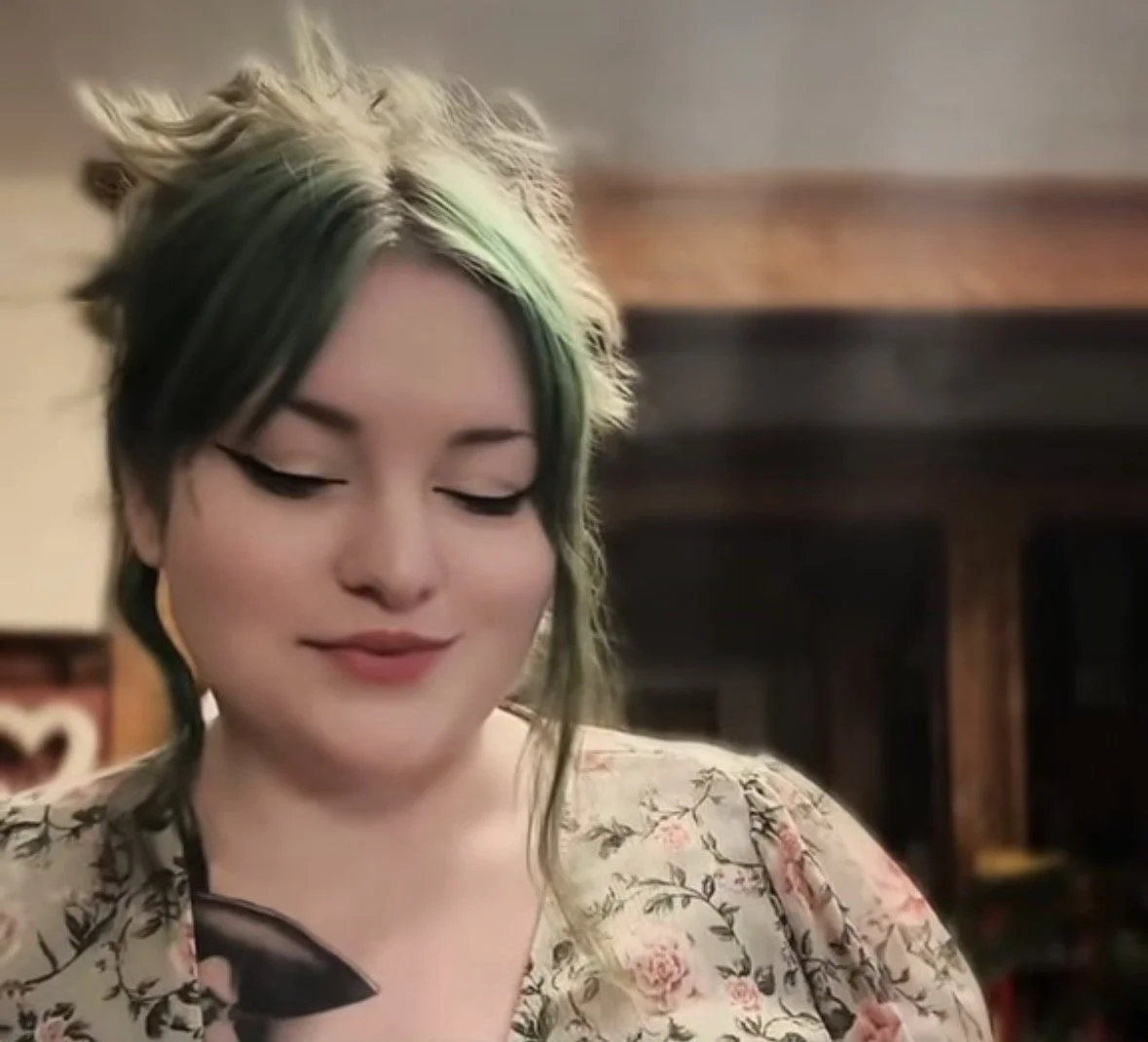 Image of Minerva Siegel, a green-haired plus size woman, from the collar bone up. She has green hair tied into a messy bun and is looking down, smiling softly.