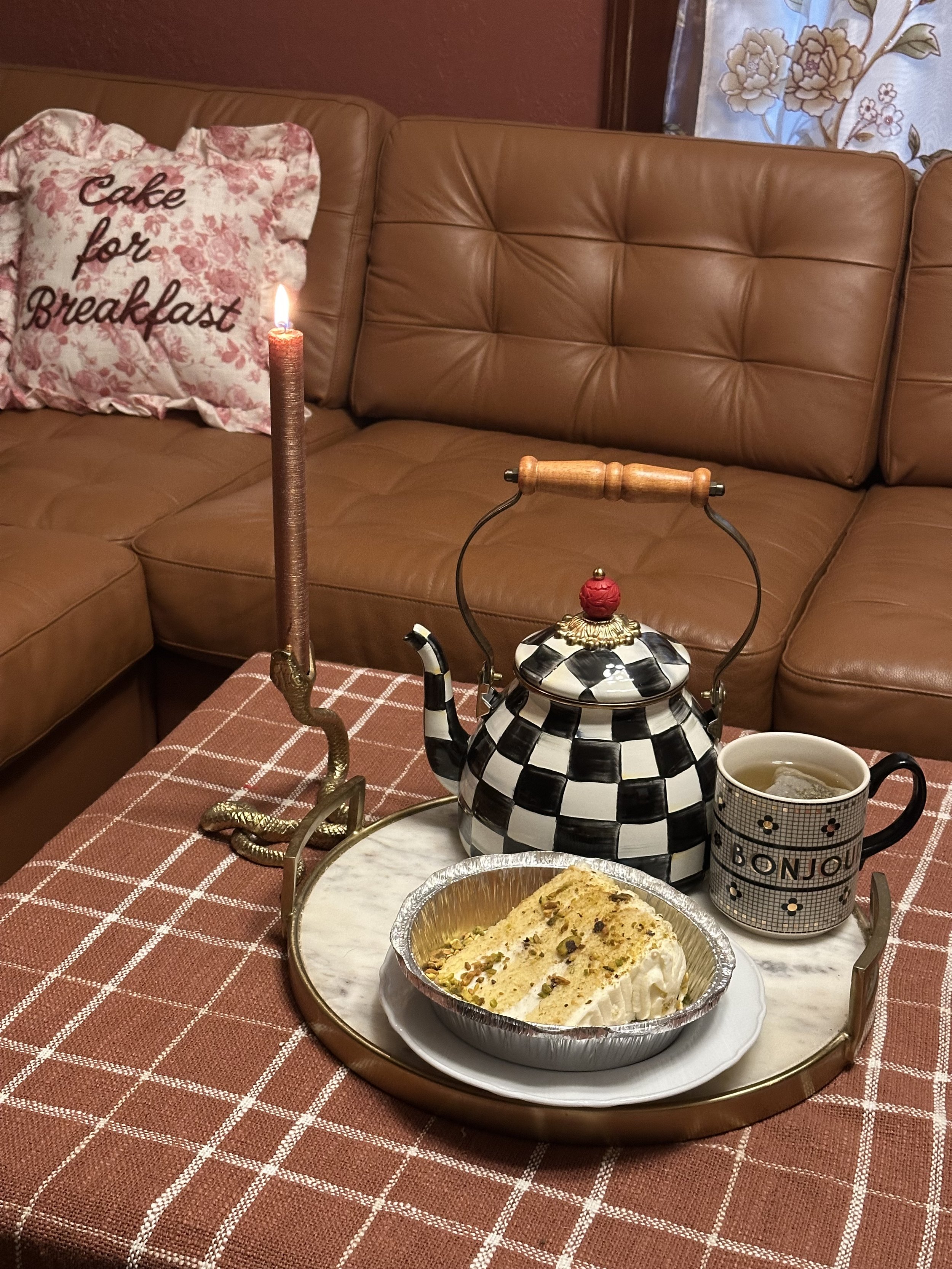 Image of a corner of a brown chesterfield sectional sofa. A checkered teapot and mug are visible lower right on the coffee table, a red and cream floral pillow sits on the sofa.