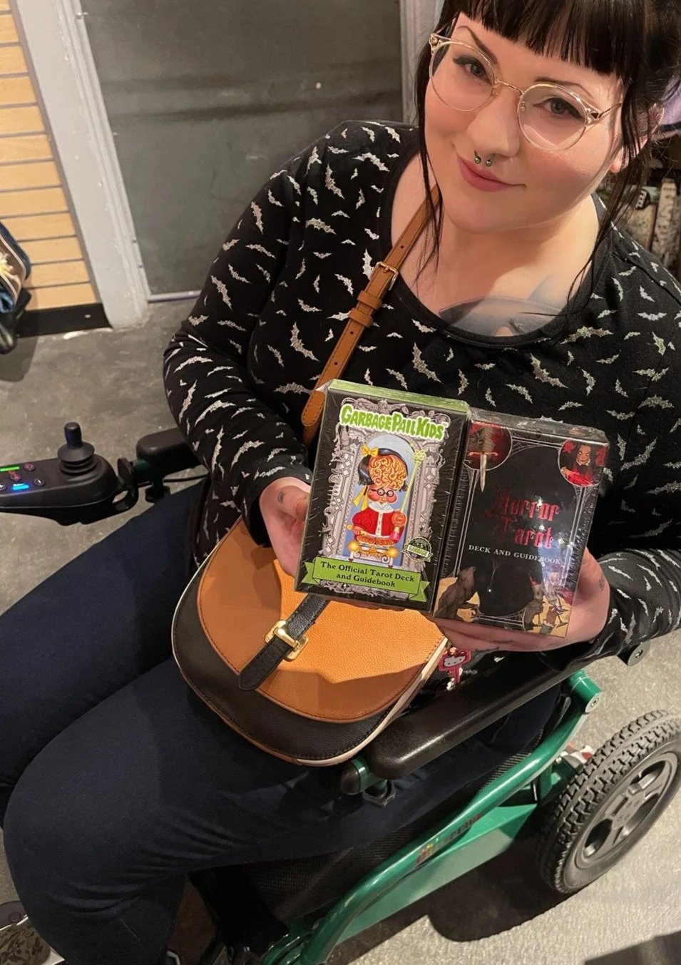 Minerva is pictured wearing a bat printed shirt and sitting in a green electric wheelchair. She's holding two tarot decks she authored and smiling in a shop where they're sold.