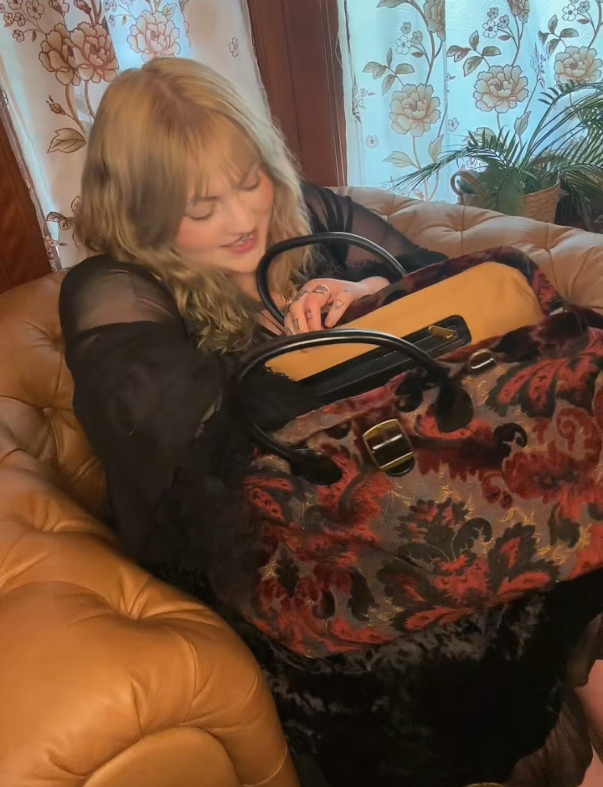 image of Minerva Siegel reaching into the Burnout Velvet Sephia Revival Max Carpetbag Works (MCW) Weekender Carpetbag. She's in her parlor on a Chesterfield chair.