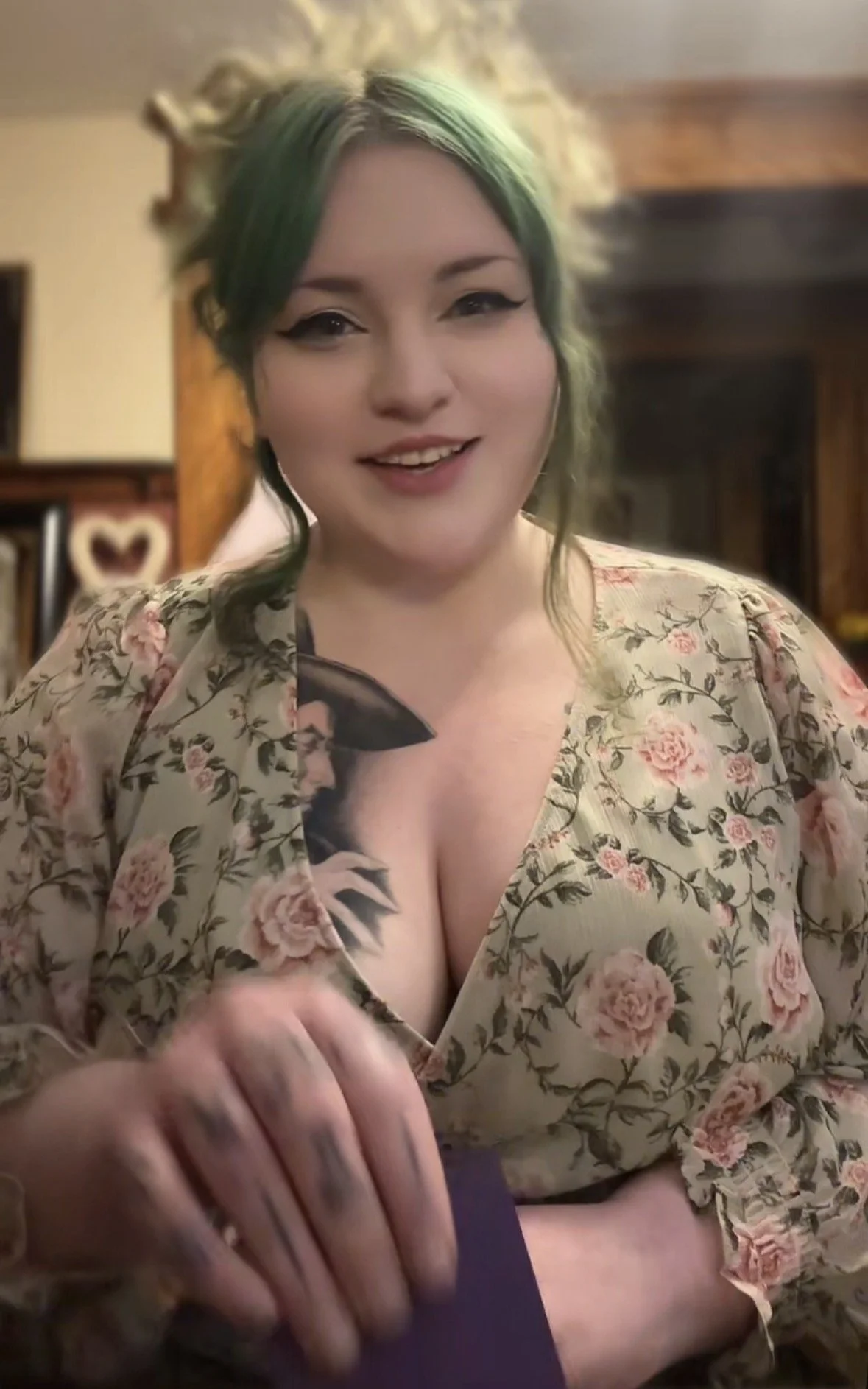 Image of Minerva Siegel during a tarot reading. She has green hair loosely tied in a messy bun, she's drawing a purple card with her tattooed fingers, and smiling warmly at the camera. She's wearing a green floral dress.