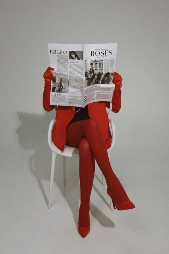 Une personne assise sur une chaise blanche, portant des vêtements rouges et des talons rouges, lisant un journal