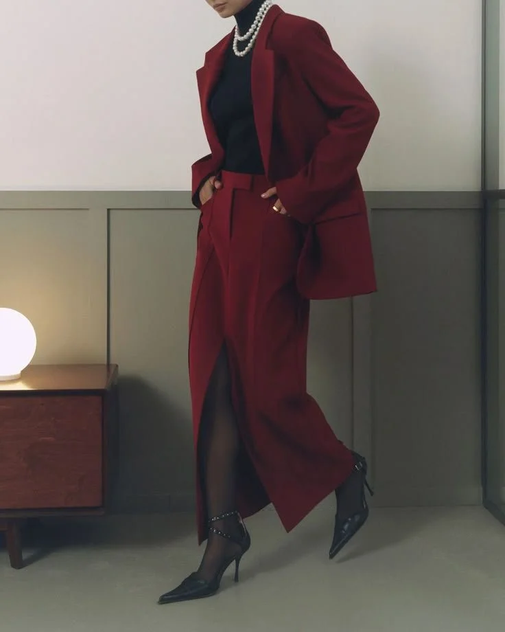 Femme portant un tailleur rouge avec une veste et une jupe longue, un haut noir, une chaîne de perles, des collants noirs et des talons hauts noirs avec des décorations.