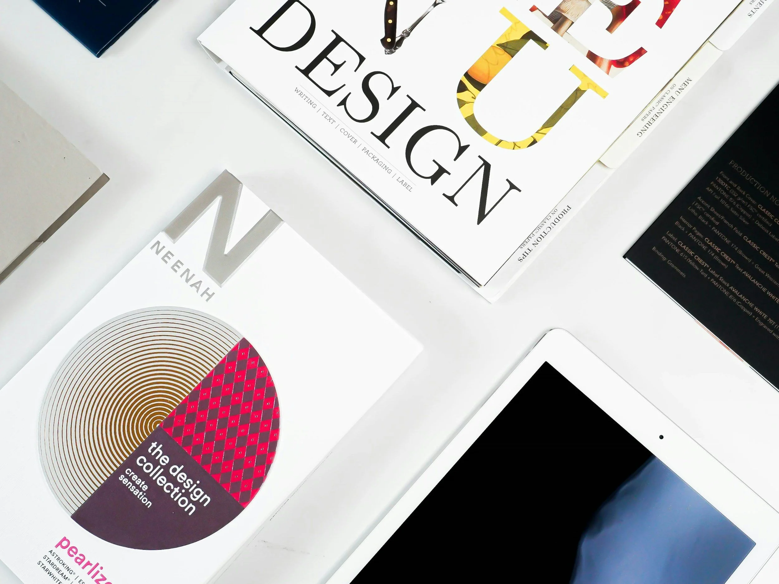 A flat lay of design books and a tablet on a white surface. The visible books include 'Design' with a colorful floral and yellow pattern on the cover, and 'Neenah' with a white cover and a circular graphic design.