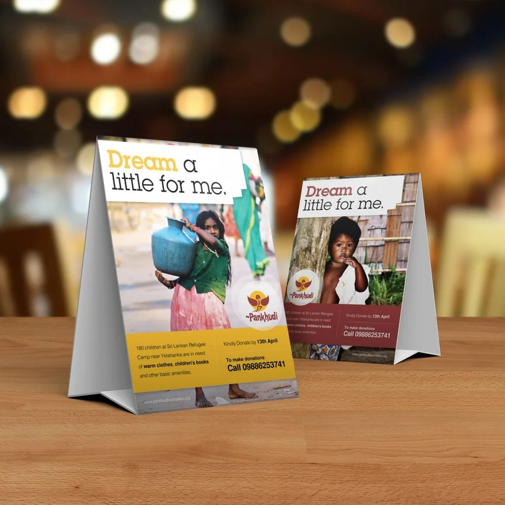Two folded charity flyers on a wooden table promoting donations for Sri Lankan refugee children, one showing a girl carrying a water container and the other a young boy.