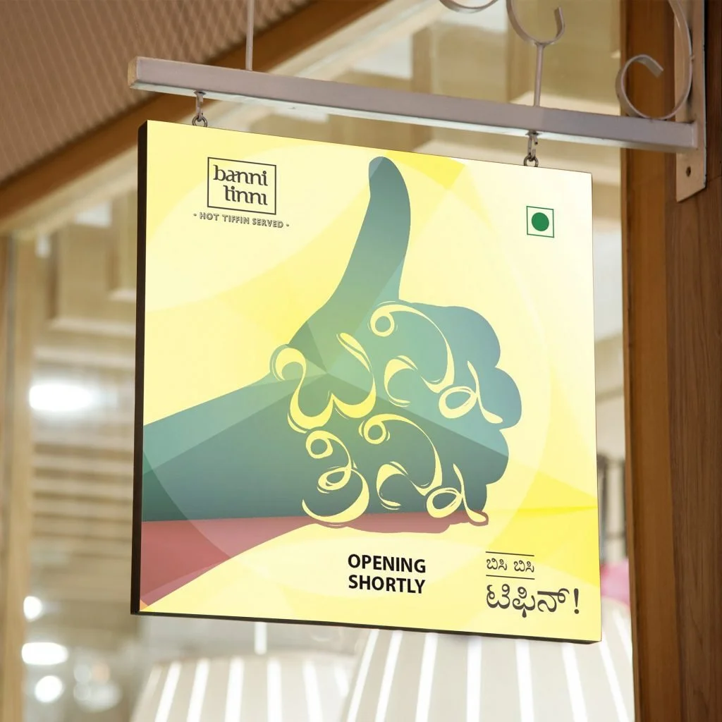 A colorful sign with abstract art and text in both English and another language. The sign reads 'banni timmi' at the top, 'HOT TIFIN SERVED' below that, and 'OPENING SHORTLY' in the middle. There is a small green square in the upper right corner.