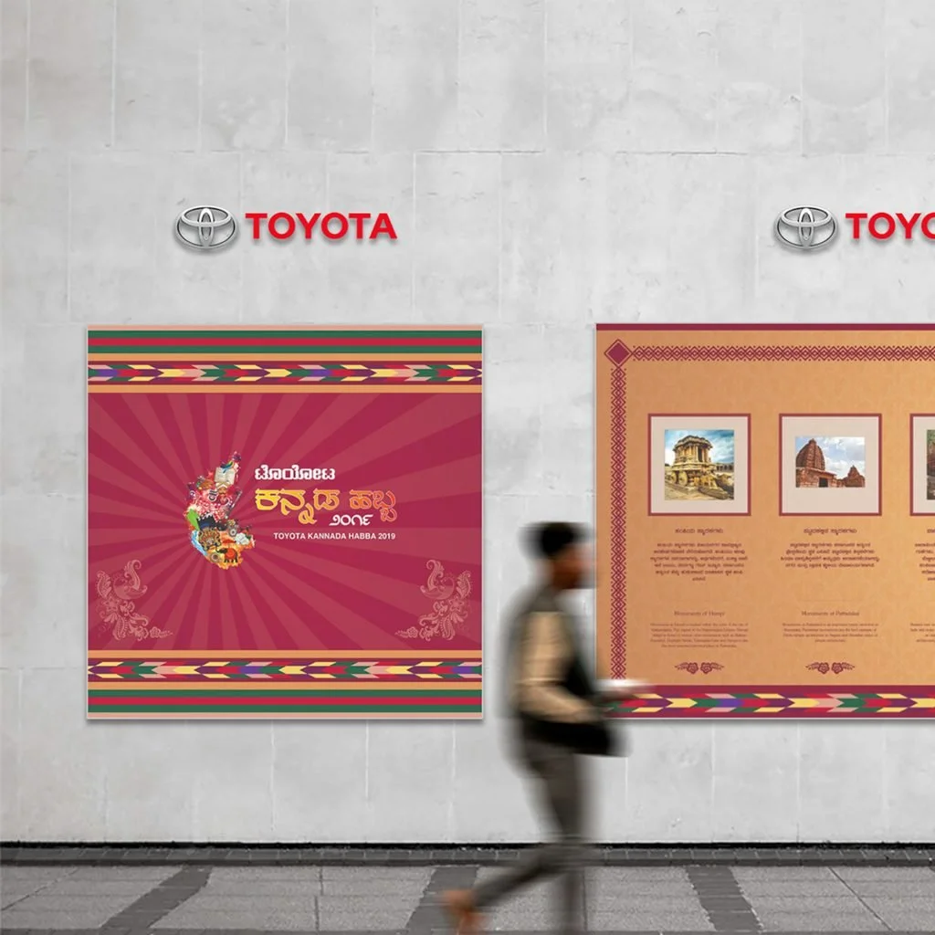 A man walking in front of a wall with two posters and the Toyota logo. The left poster features a colorful design and text in Kannada, celebrating the Toyota Kannada Habba 2019. The right poster has images of ancient temples and descriptions in Kannada.