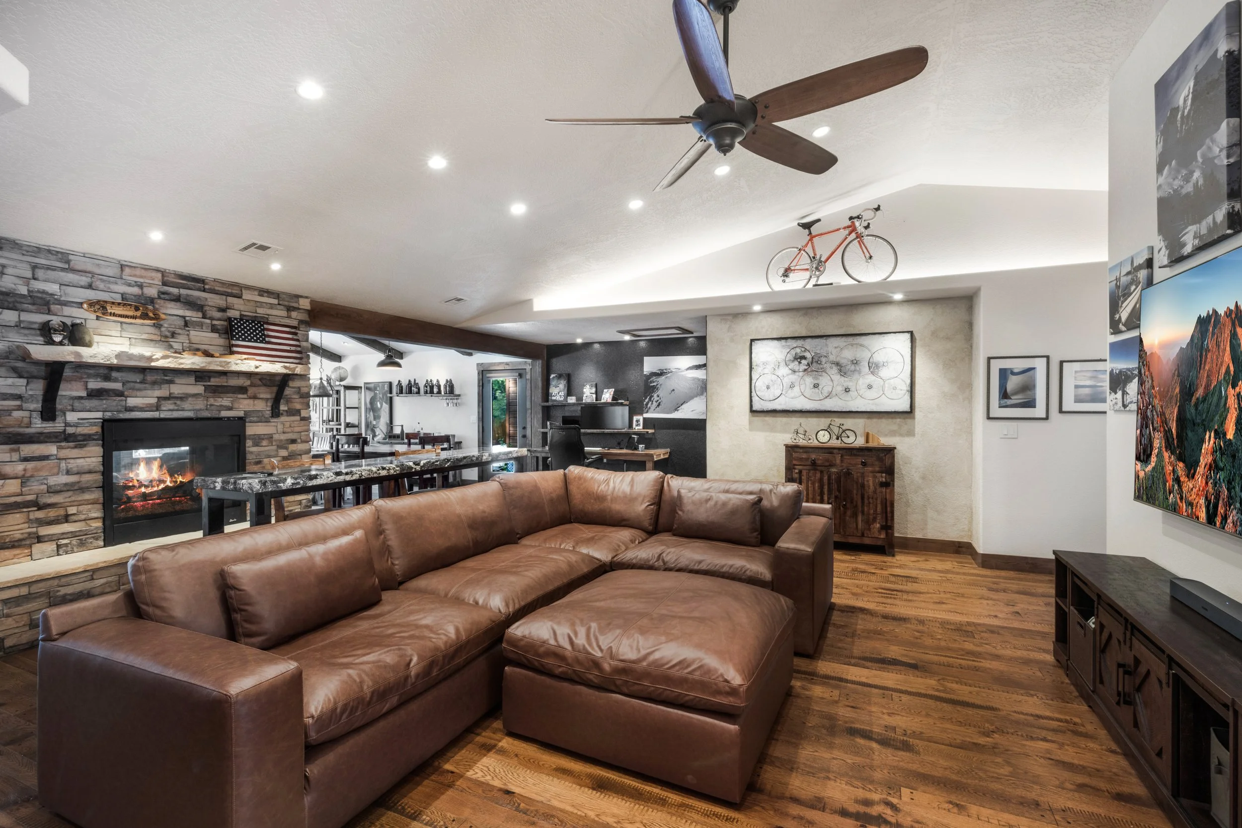 A living room with a large brown leather sectional sofa, a stone fireplace with a fire, a mounted ceiling fan, a wooden TV stand with a flat-screen TV, and various bicycle artwork and decorations on the walls, with a dining area in the background.