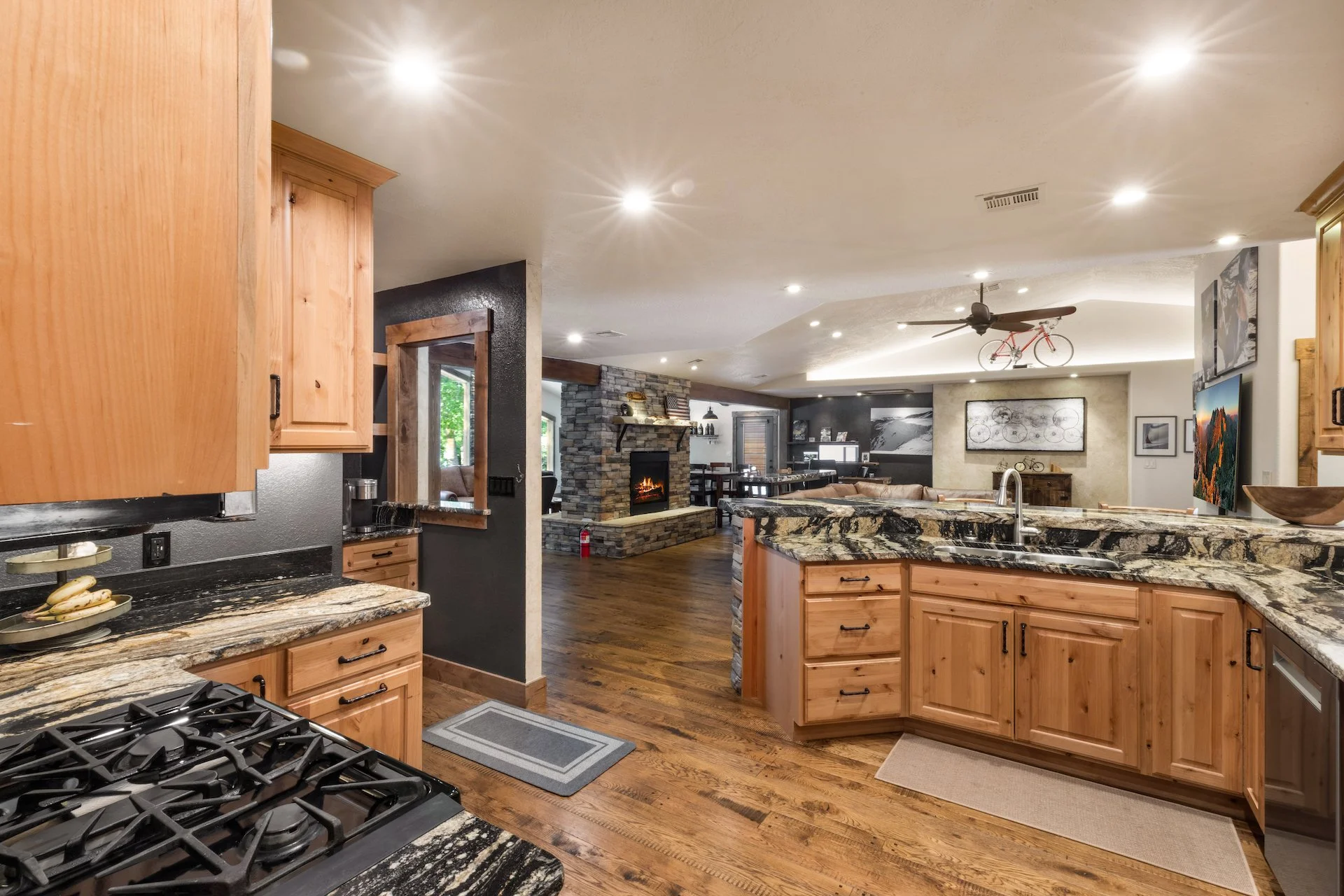 Open-concept kitchen and living room with wooden cabinets, granite countertops, fireplace, hardwood floors, and artwork on the walls.