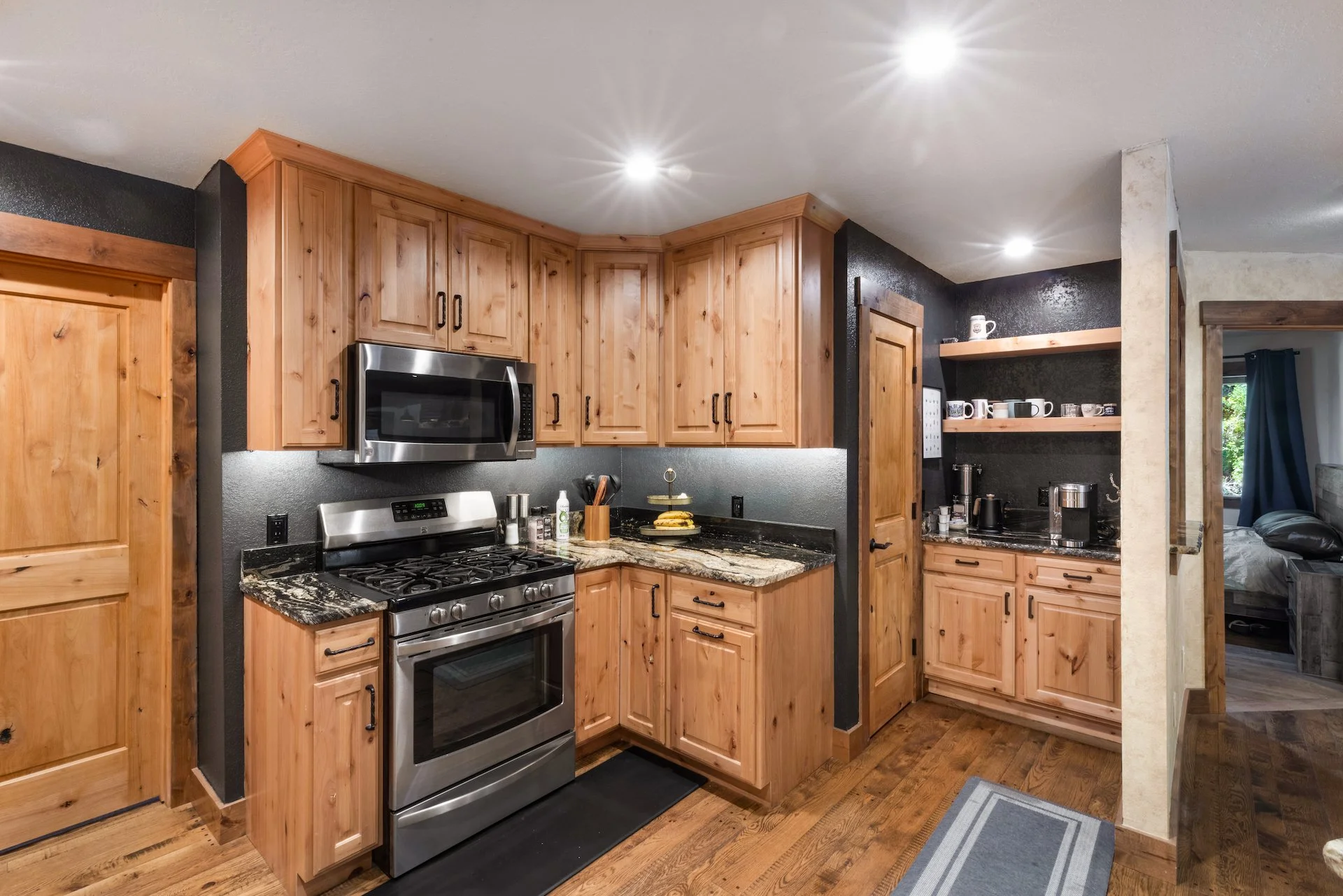 Kitchen with wooden cabinets, stainless steel stove and microwave, granite countertops, and open shelving with cups and coffee-making equipment.