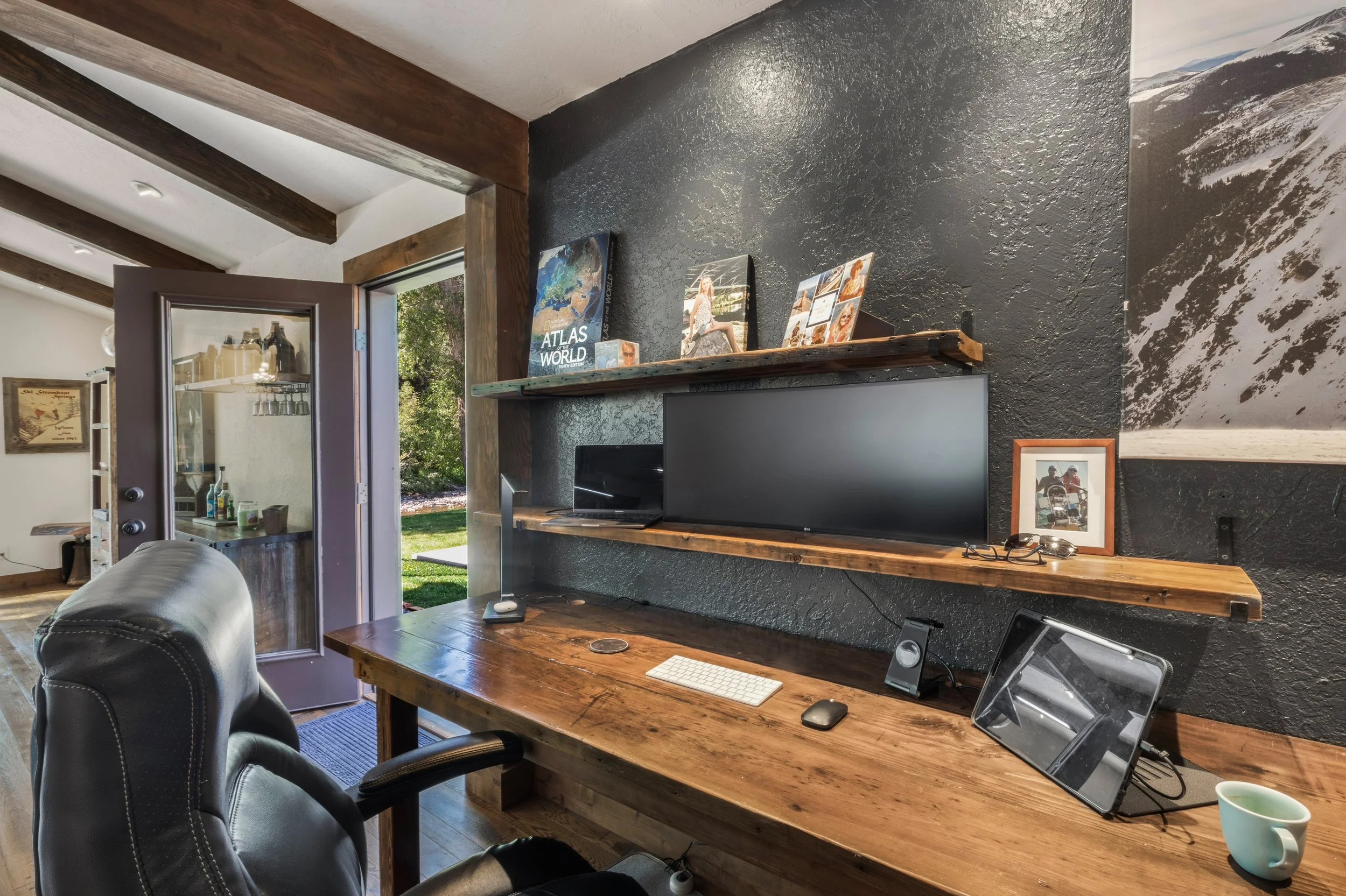 Home office setup with a wooden desk, computer monitor, tablet, and office chair, black textured wall with shelves holding books and photographs, open door leading to a green backyard.