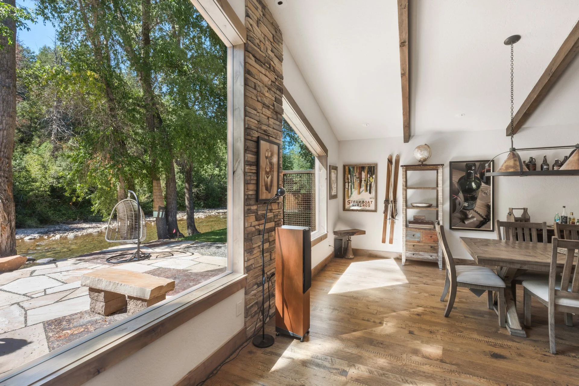 Interior view of a dining and living space with large windows showing outdoor trees and a river, wooden furniture, exposed beams, and artwork on the walls.