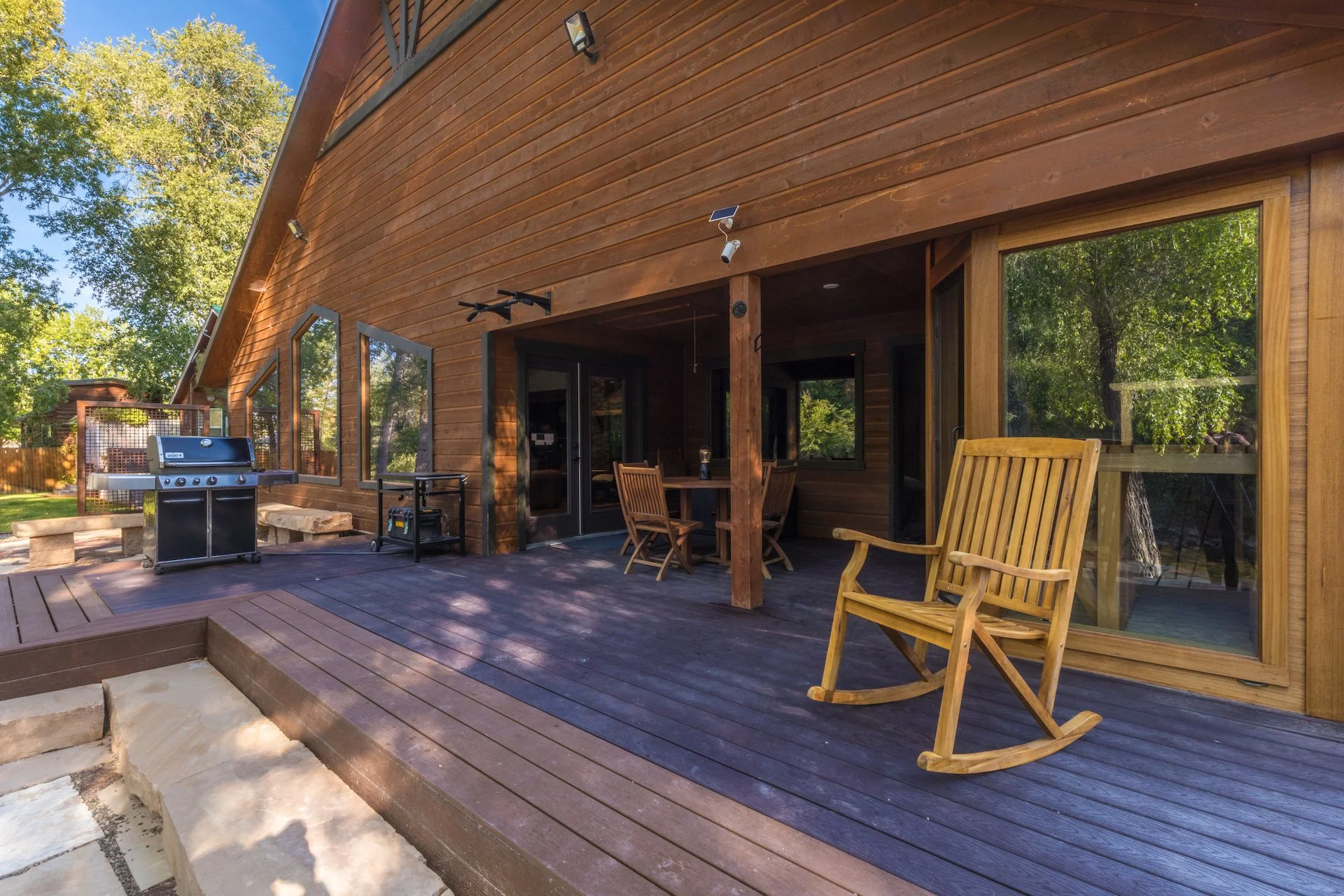 Wooden patio with a rocking chair, a round table with chairs, a barbecue grill, and large windows, surrounded by trees.