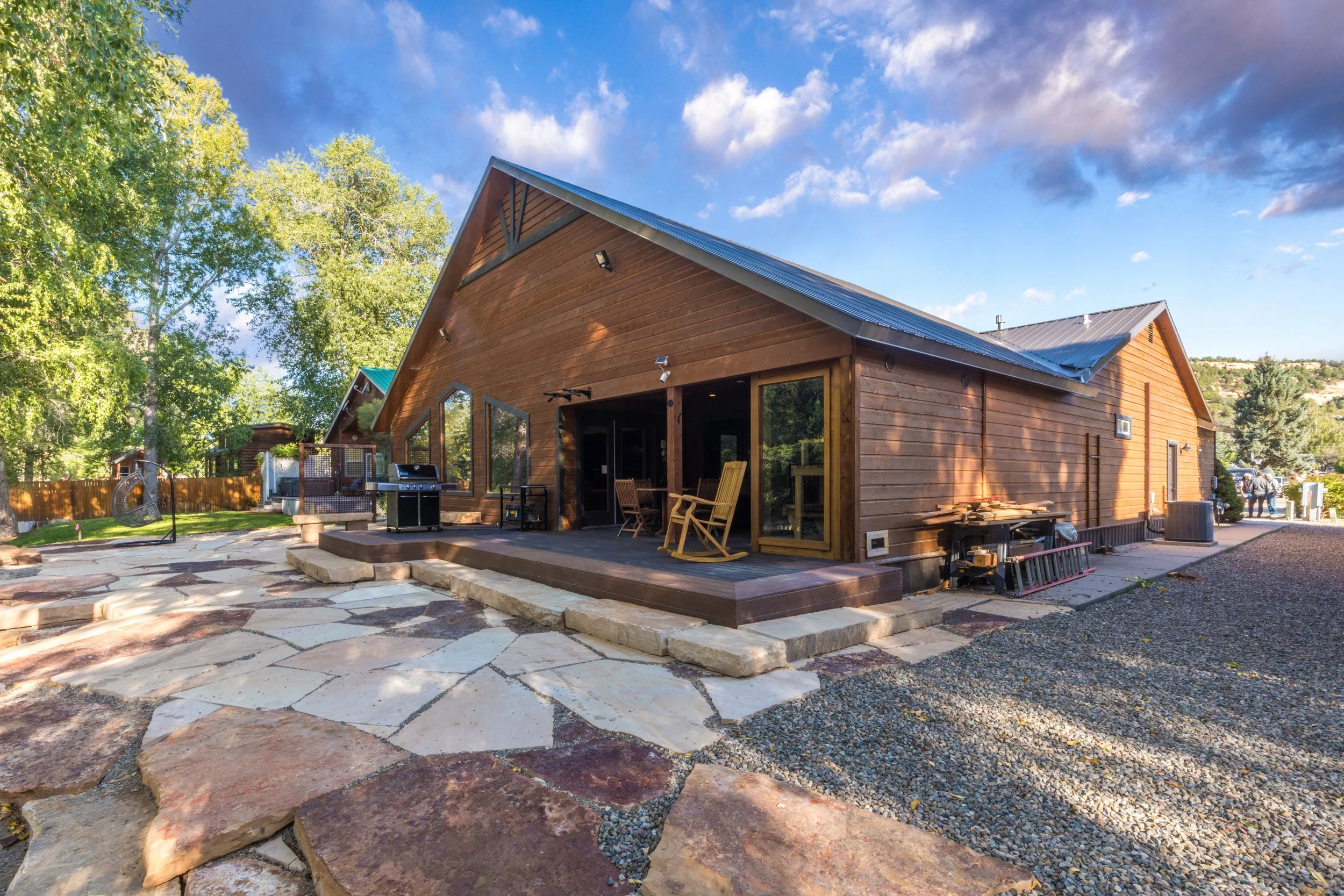 A wooden house with a large deck, outdoor furniture, and a grill, surrounded by trees and a stone patio, under a partly cloudy sky.
